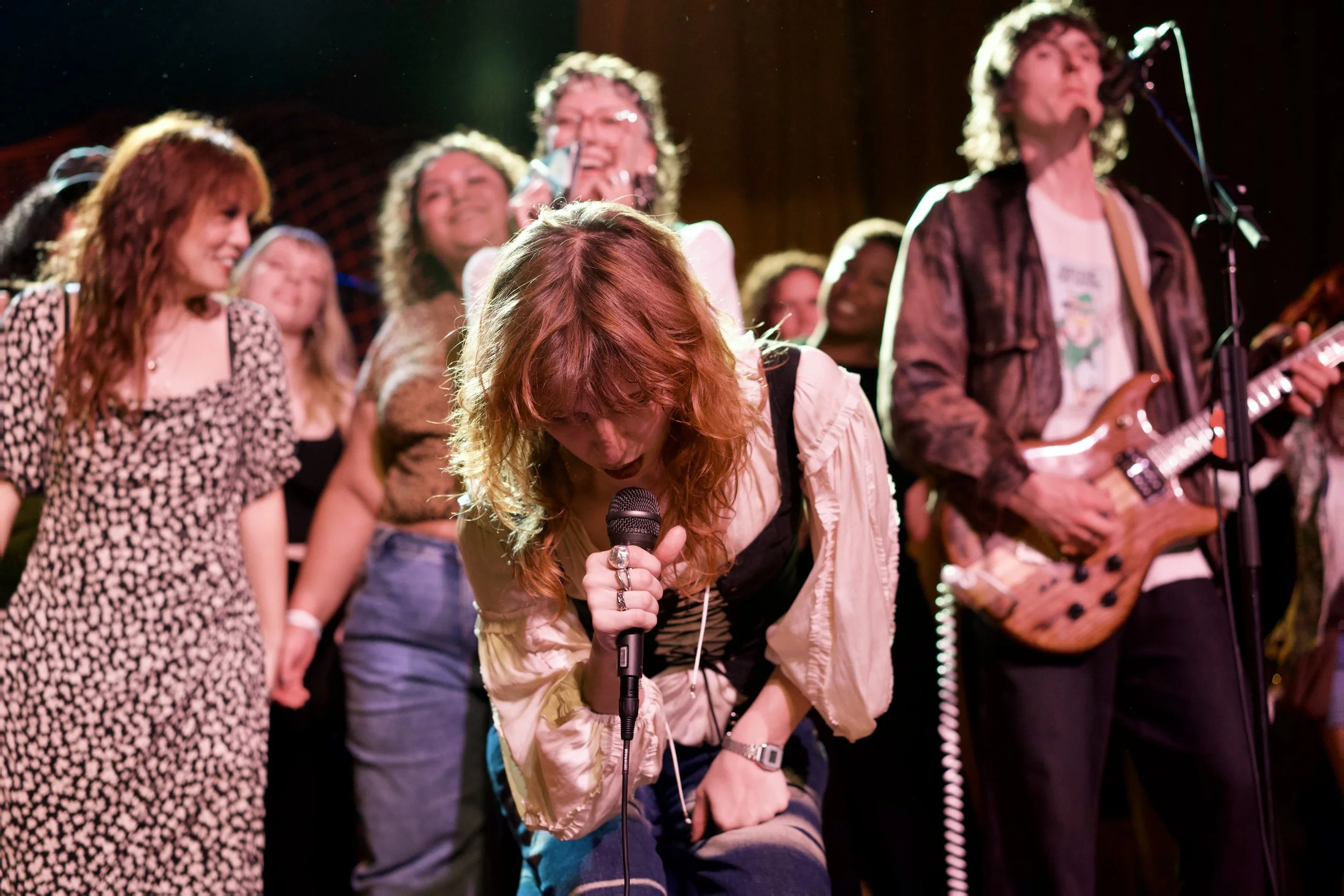 Group of people performing on stage, with a woman singing into a microphone in the foreground, others playing guitar and smiling, stage lights overhead.