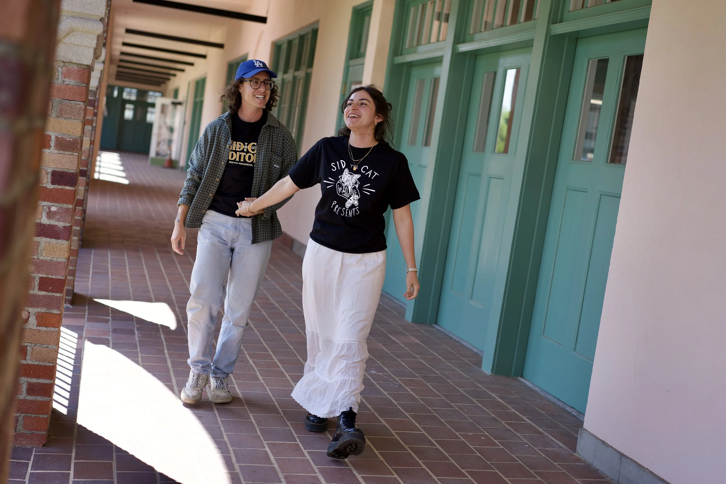Two women walking hand in hand in a corridor with green doors and brick walls, smiling and looking at each other.