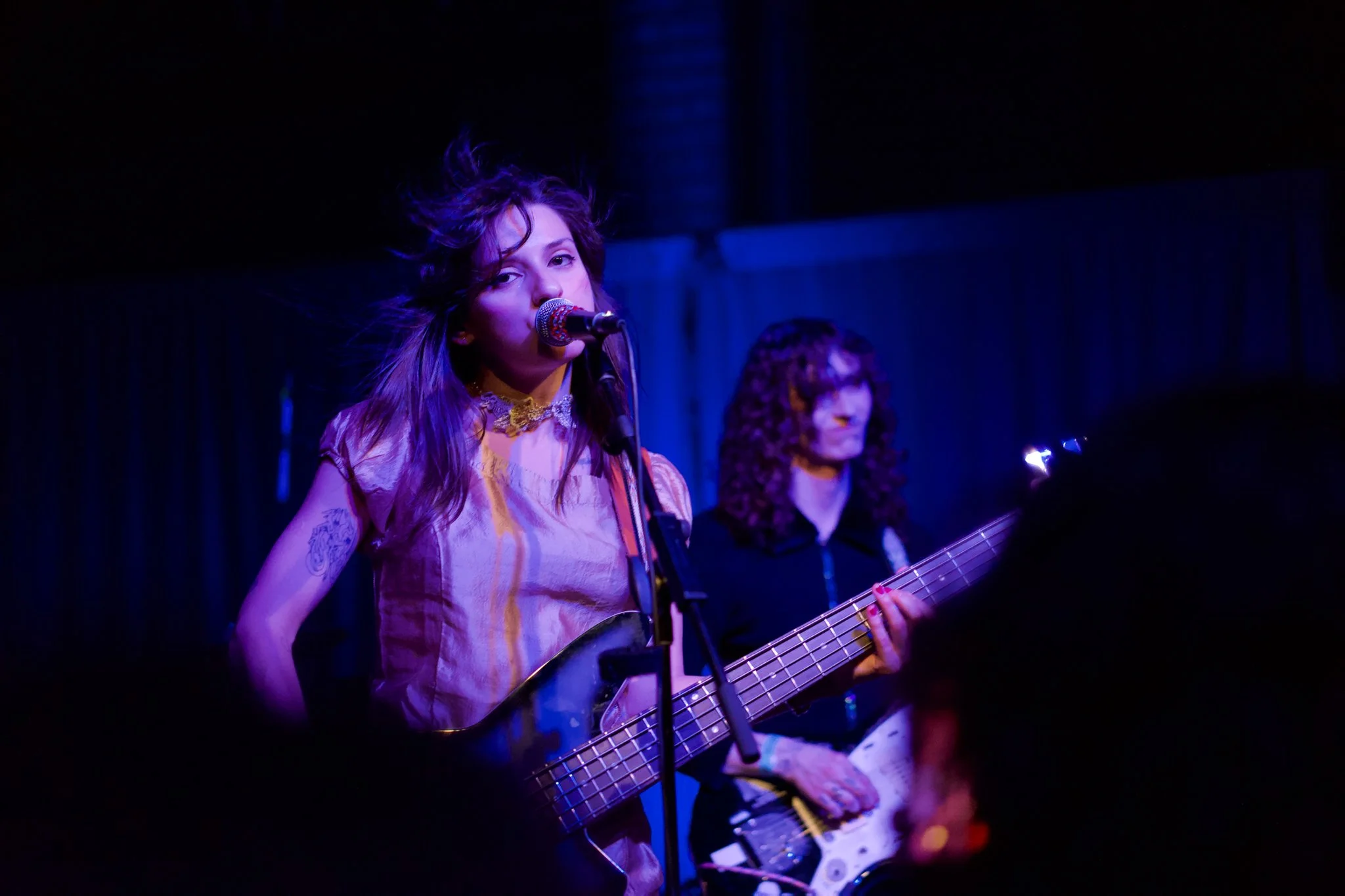A young woman with long hair singing into a microphone and playing an acoustic guitar on stage, with another person in the background playing bass guitar, both performing under dark blue and purple stage lighting.