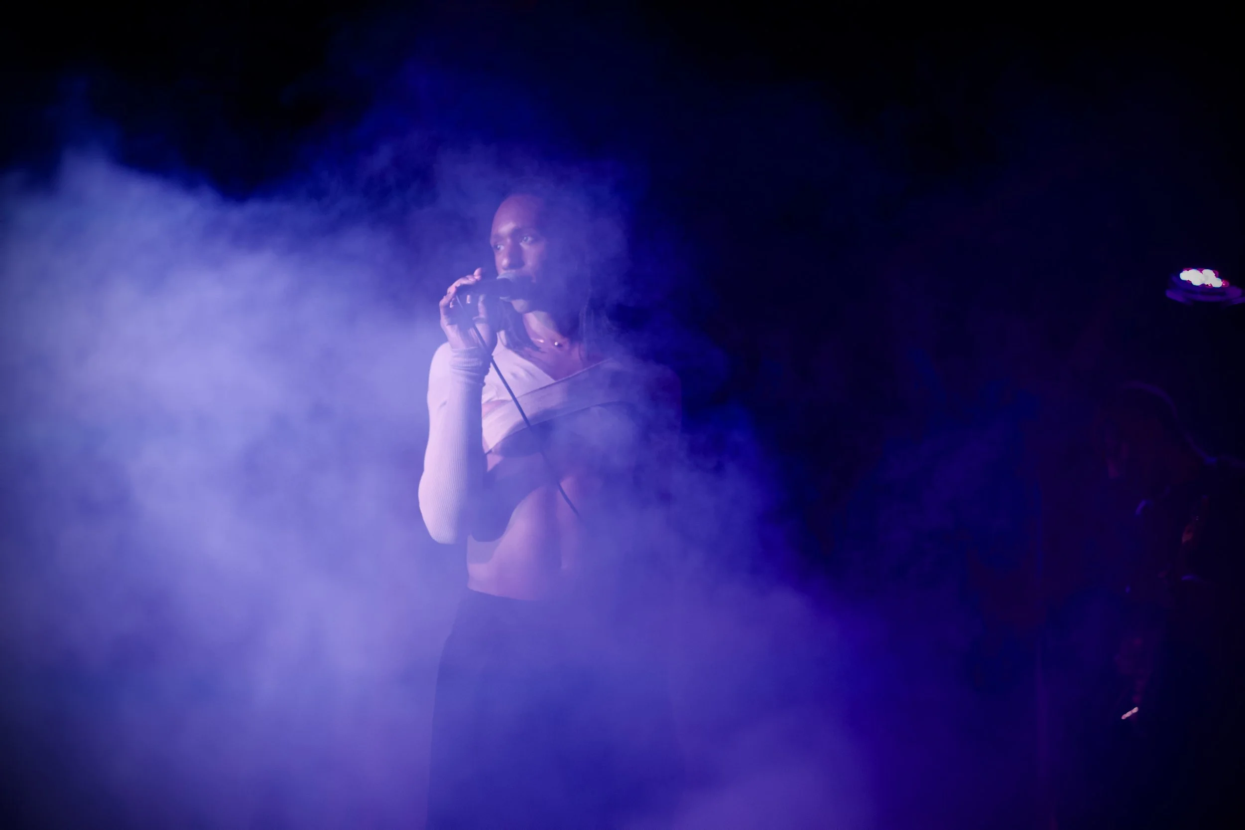 A singer with dark hair and a light-colored long sleeve shirt holding a microphone, performing in a dark setting with blue and purple stage lighting.