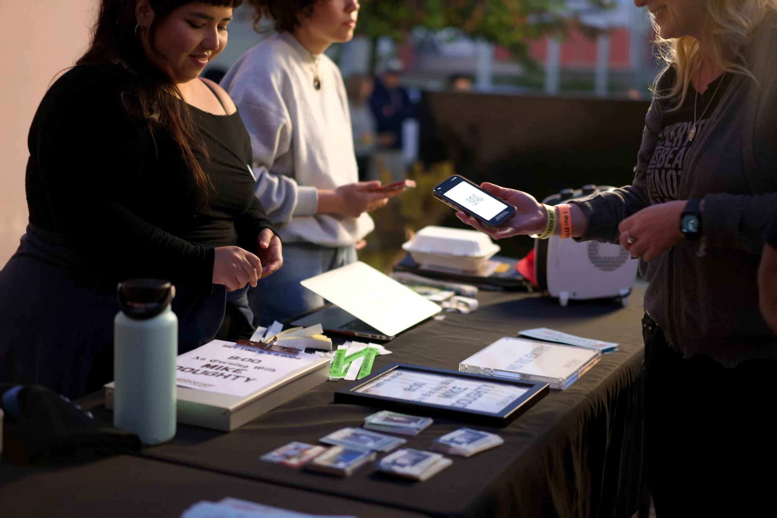 Three women standing at a table outdoors at dusk, looking at a smartphone displaying a QR code, with pamphlets and books on the table.