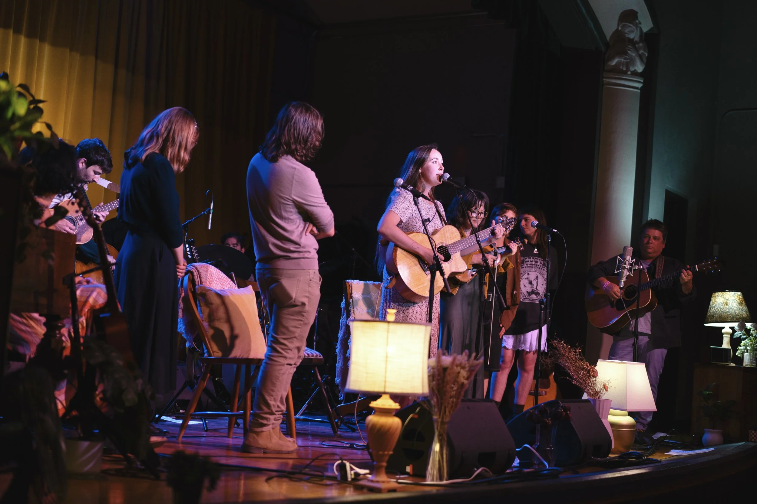 Group of people performing music on stage with guitars and microphones, illuminated by warm stage lights, with some standing and others sitting, in a cozy indoor setting.