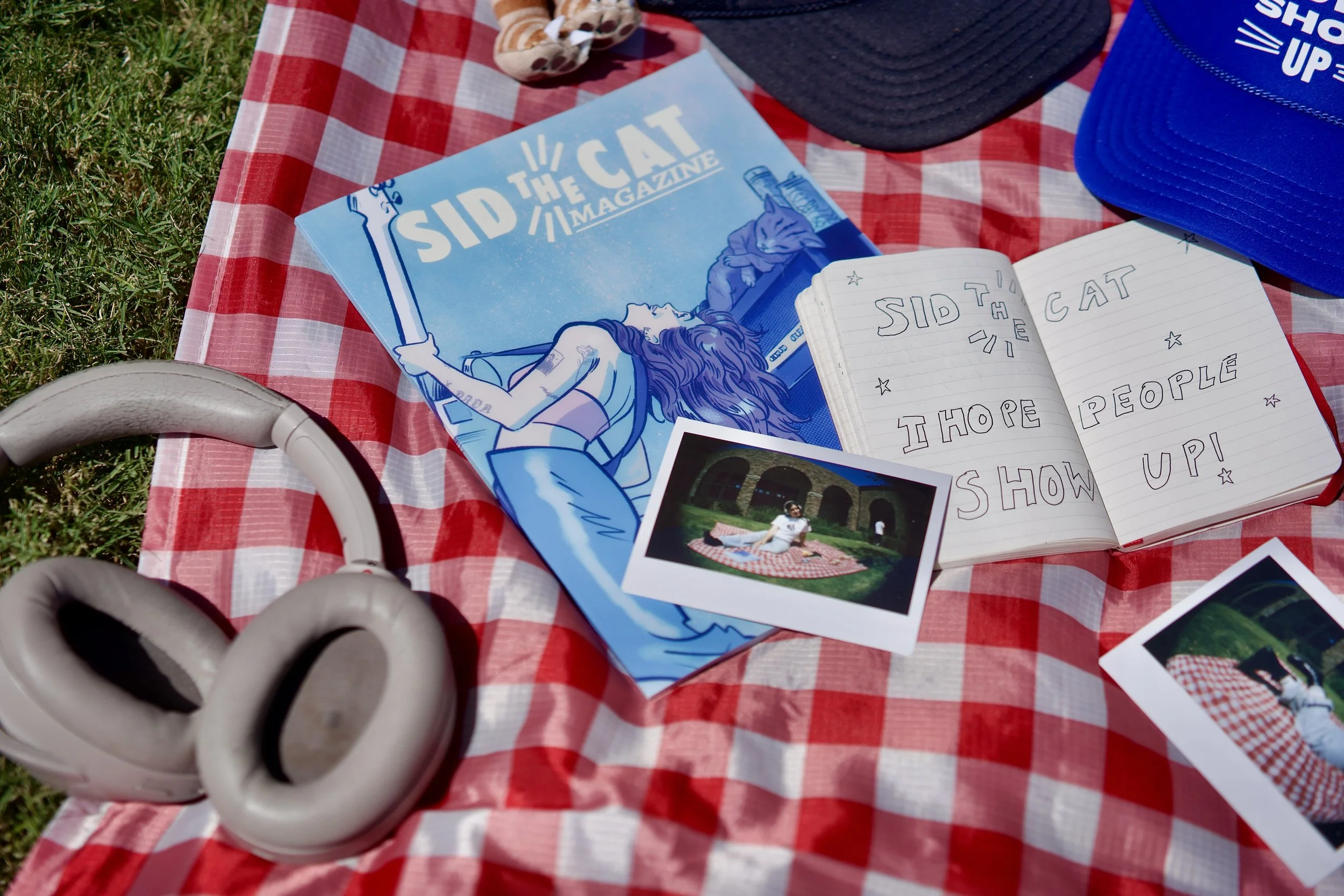 Picnic setup on a red and white checkered blanket with a baseball cap, a magazine titled 'Sid the Cat' with blue cover art, a notebook with handwritten message, a photo of a person on a picnic blanket, and a pair of white over-ear headphones.