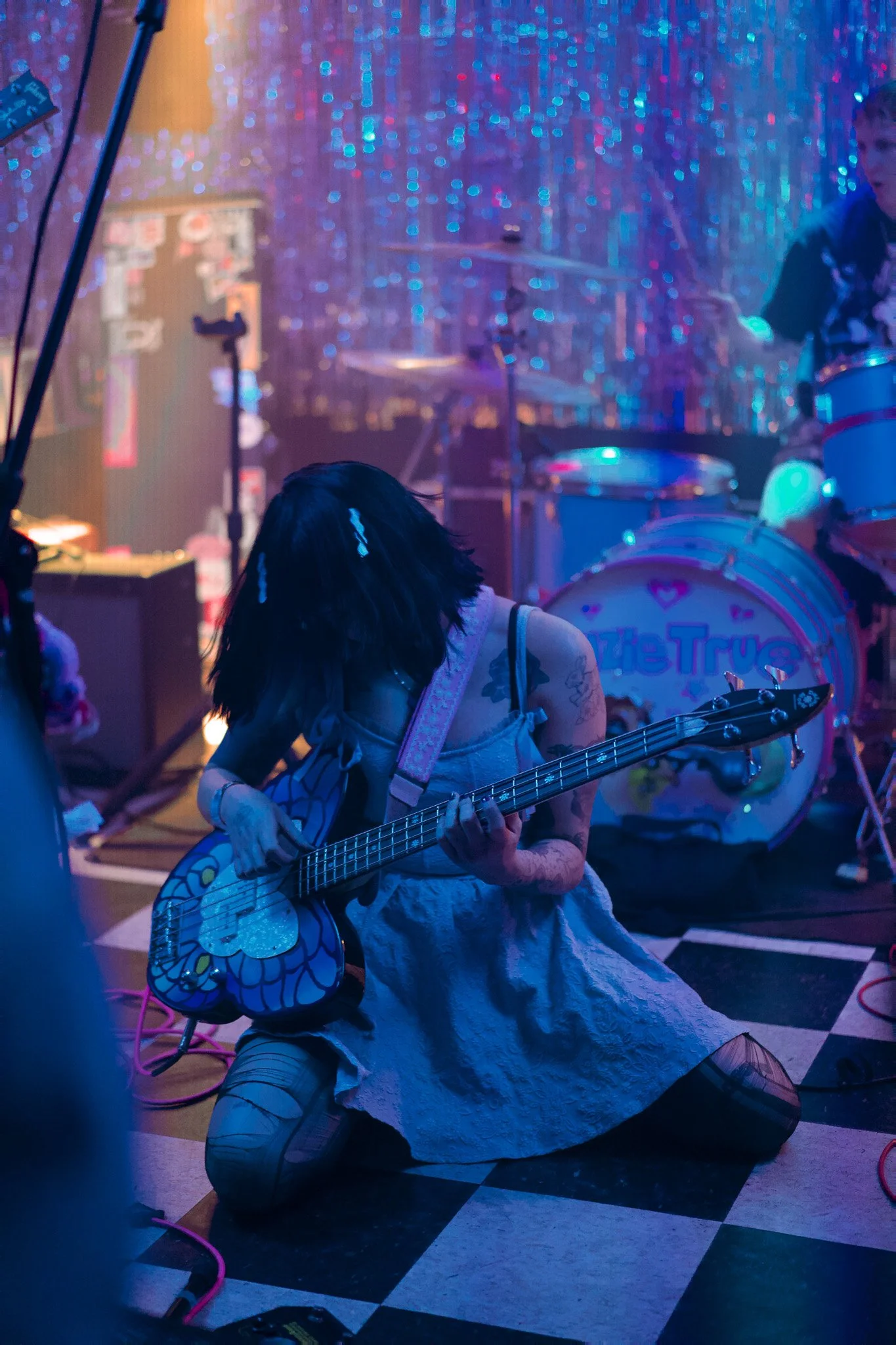 A woman kneeling on the floor playing an electric guitar with a colorful, patterned body. She is in a room with a shiny, iridescent backdrop and is dressed in a long dress with tattoos visible on her arms. A man in a dark shirt is playing drums in the background. The scene is lit with multicolored stage lighting.
