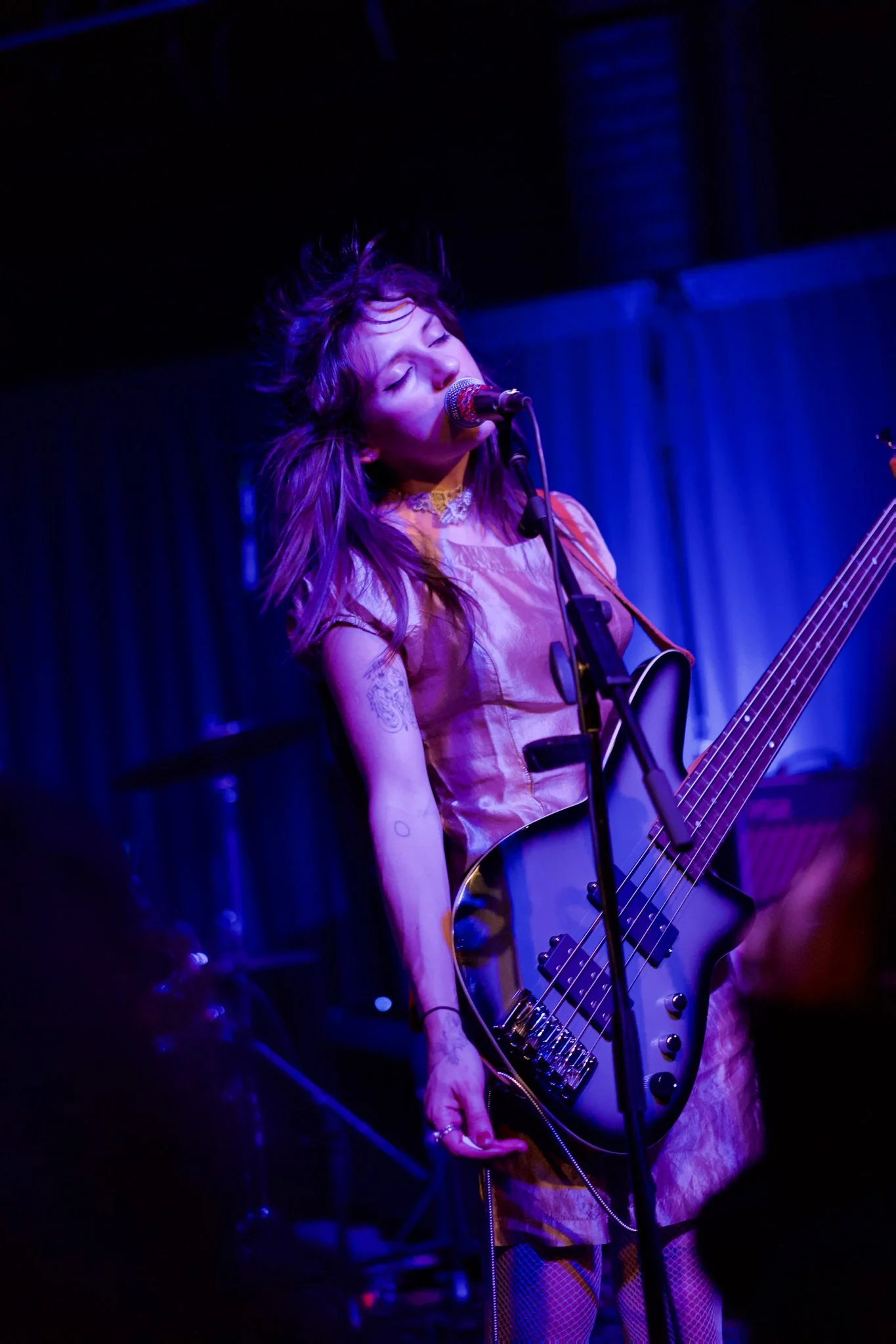 A female musician singing into a microphone and playing an electric guitar on stage, with her eyes closed and hair tousled, under blue stage lighting in a dark venue.