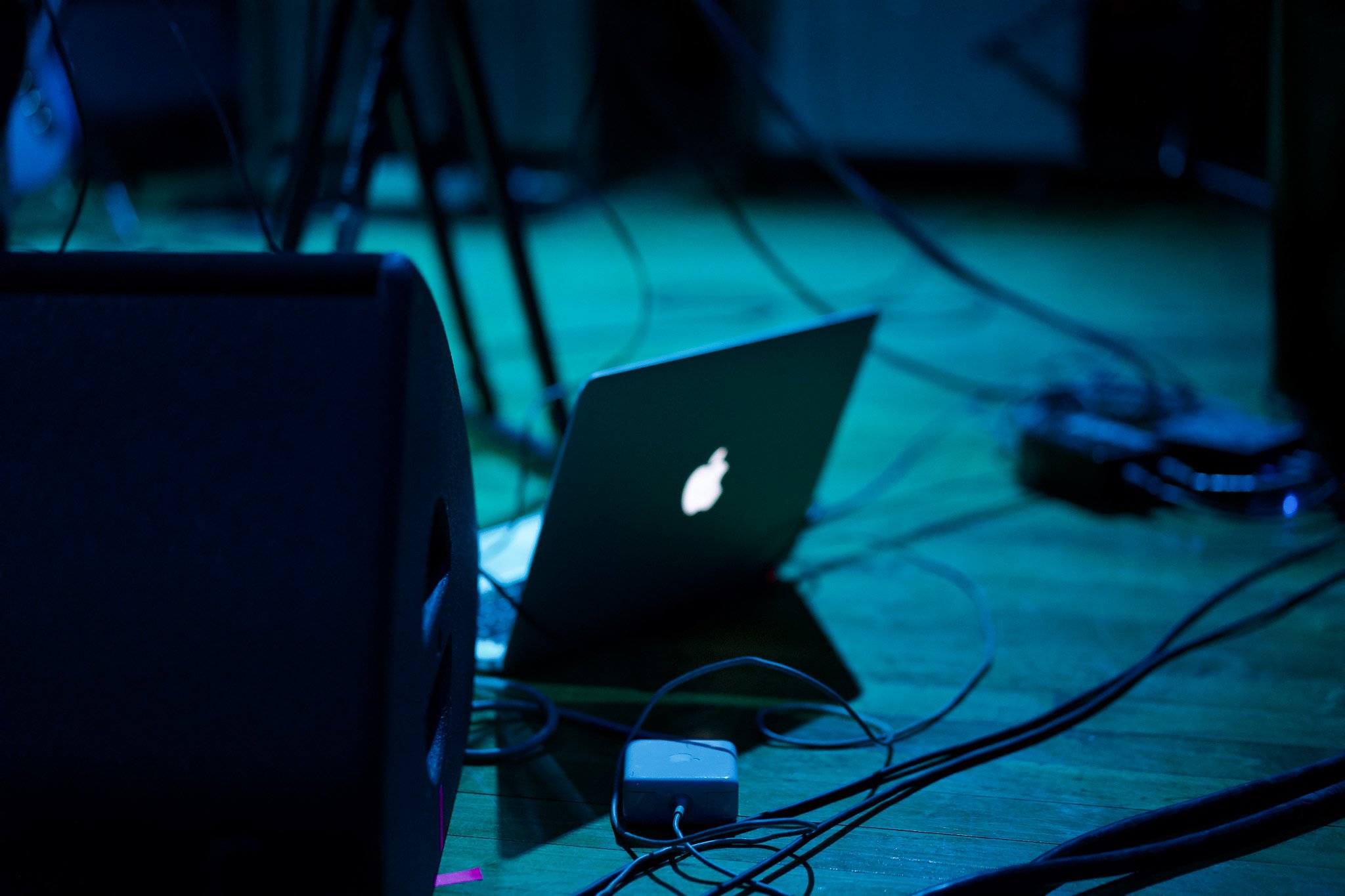 Close-up view of electronic equipment including cables, power strips, and a laptop, with a blue glow, on a wooden surface.