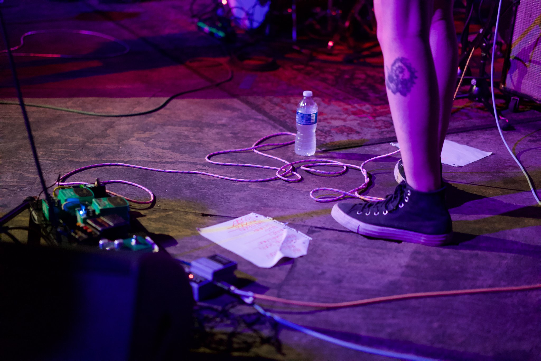 A person's legs and feet wearing black sneakers with white soles, on a dark wooden stage floor with colorful lighting. There are various cables, guitar pedals, a water bottle, and a sheet of paper with handwritten notes on the floor, indicating a live music performance or rehearsal.