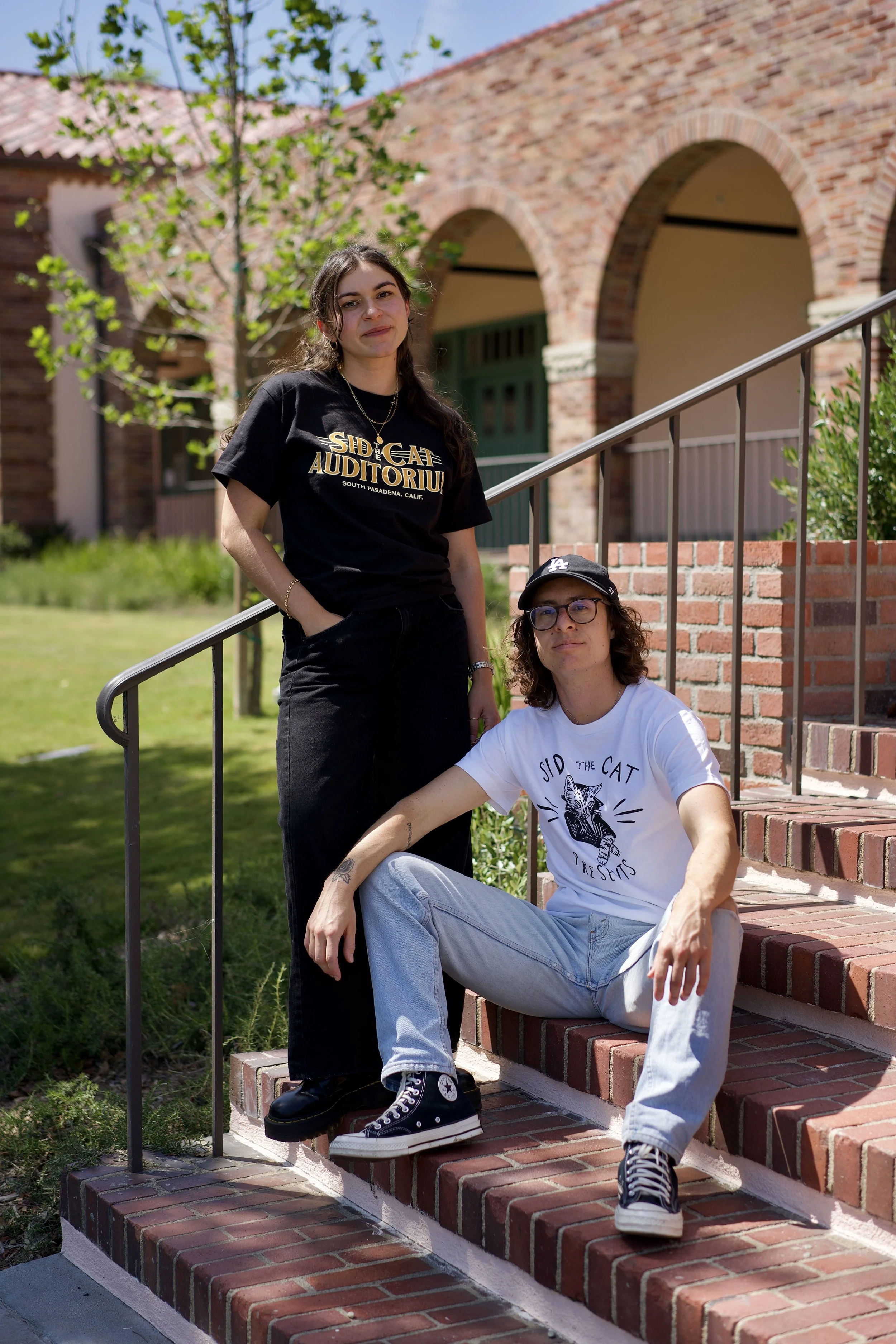 Two young adults on brick stairs outside a building. One seated on stairs wearing a white T-shirt, jeans, and a black cap; the other standing on stairs wearing a black T-shirt. The background features green trees and a brick building with arches.