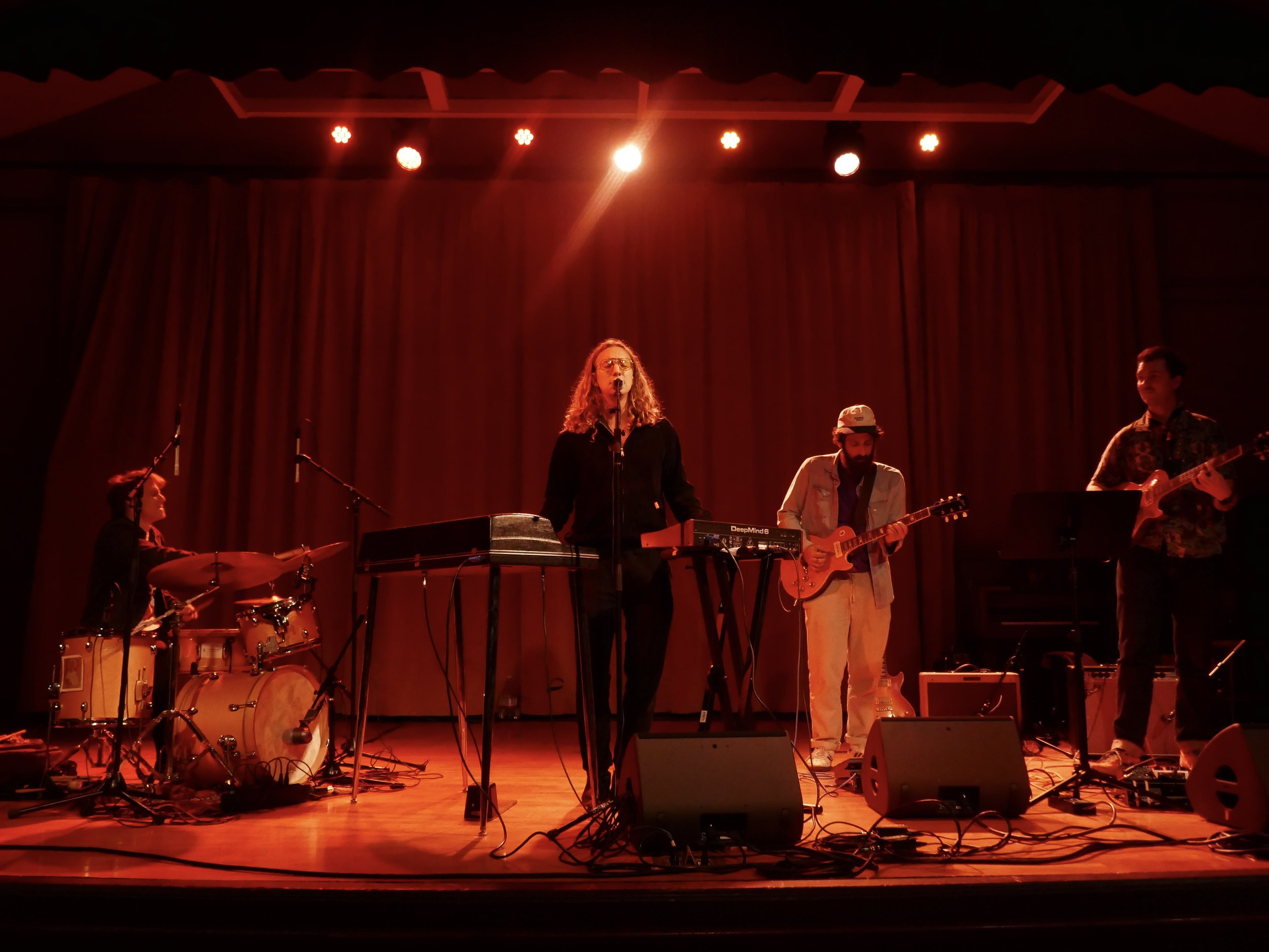 A band performs on a stage with red lighting and a red curtain backdrop, featuring a female singer, a guitarist, a keyboardist, a drummer, and another guitarist or bassist.
