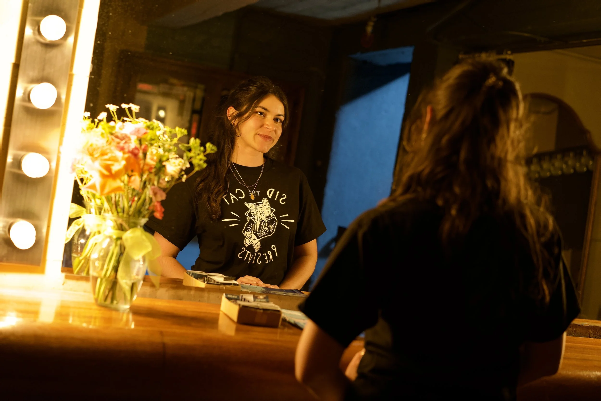 A woman with long dark hair smiling at another person in a dimly lit dressing room or backstage area, with a large mirror and lightbulbs around it, and a bouquet of colorful flowers on the table.