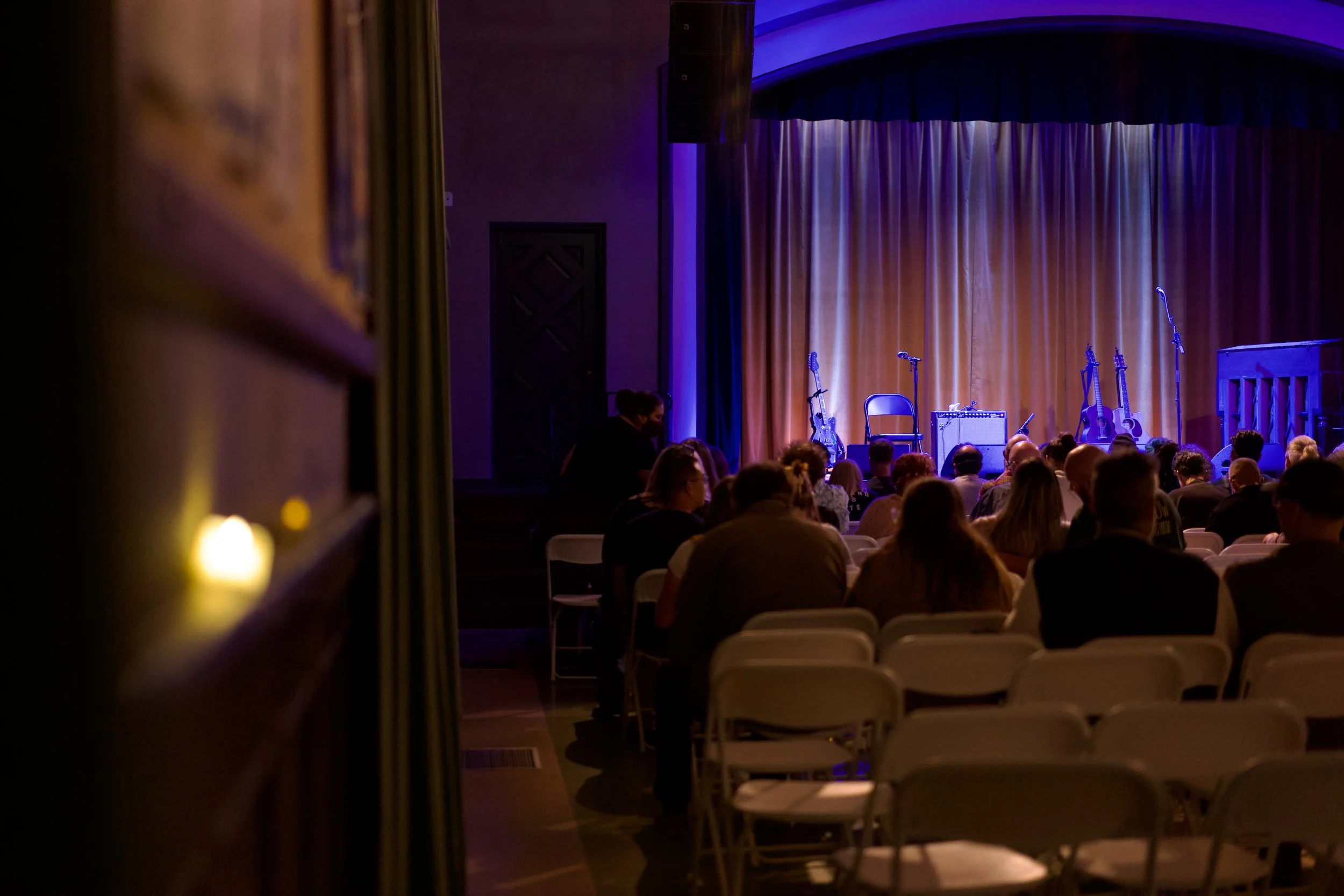 An empty stage with microphones, guitars, and amplifier, set for a musical performance, viewed from audience seating.
