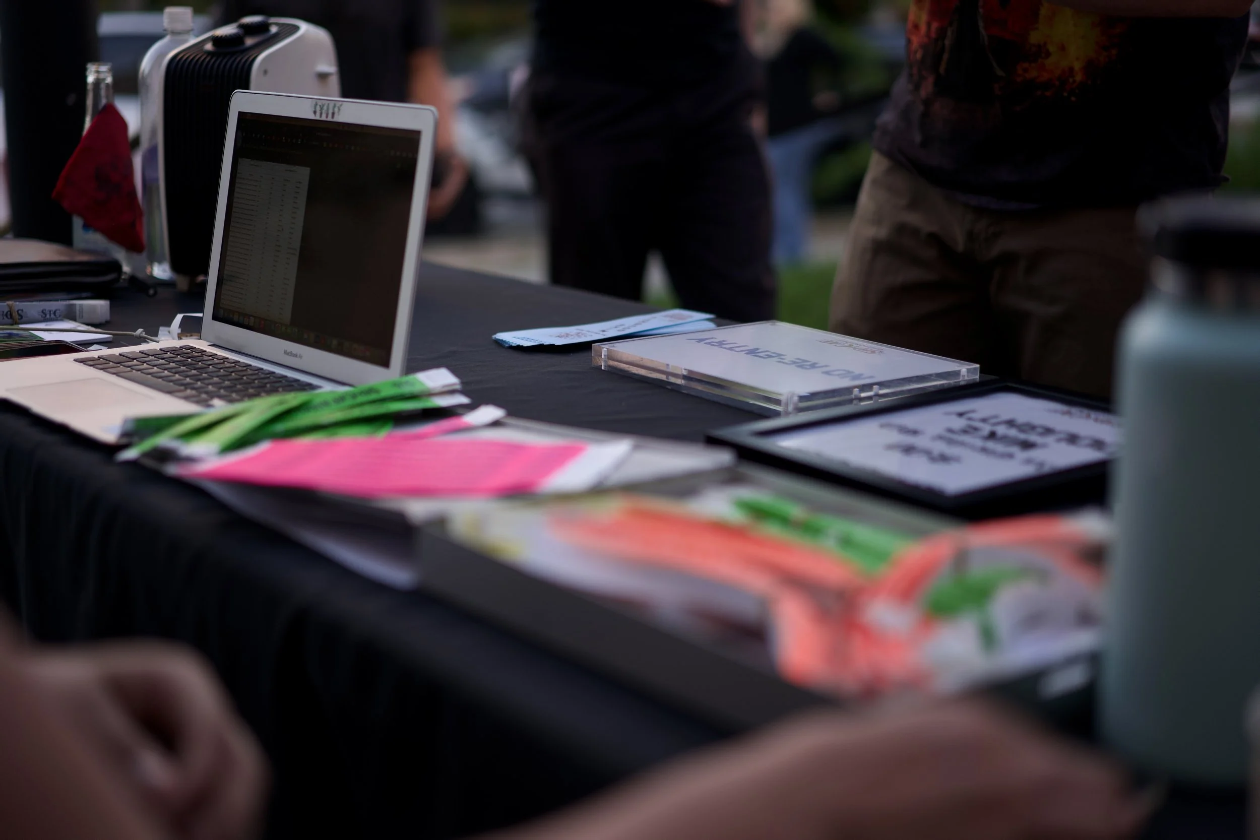 A table with various promotional materials, a laptop displaying a spreadsheet, a CD case, and colorful flyers, set up outdoors during daytime.