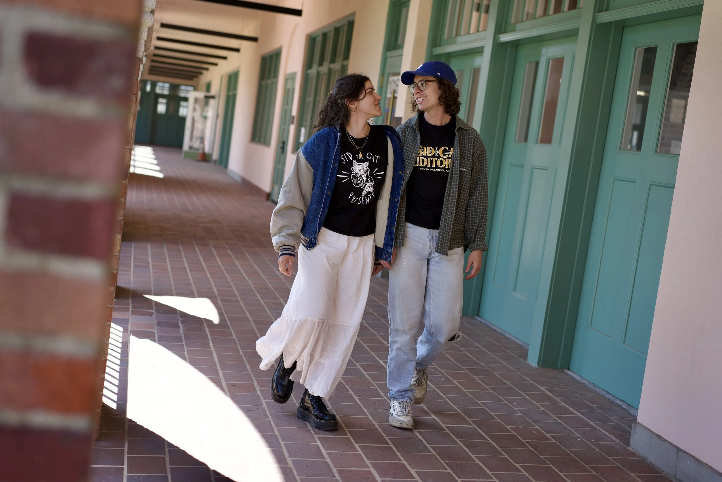 Two young people walking and smiling at each other in a corridor with green doors and windows, brick floor, and brick wall partially visible on the left.