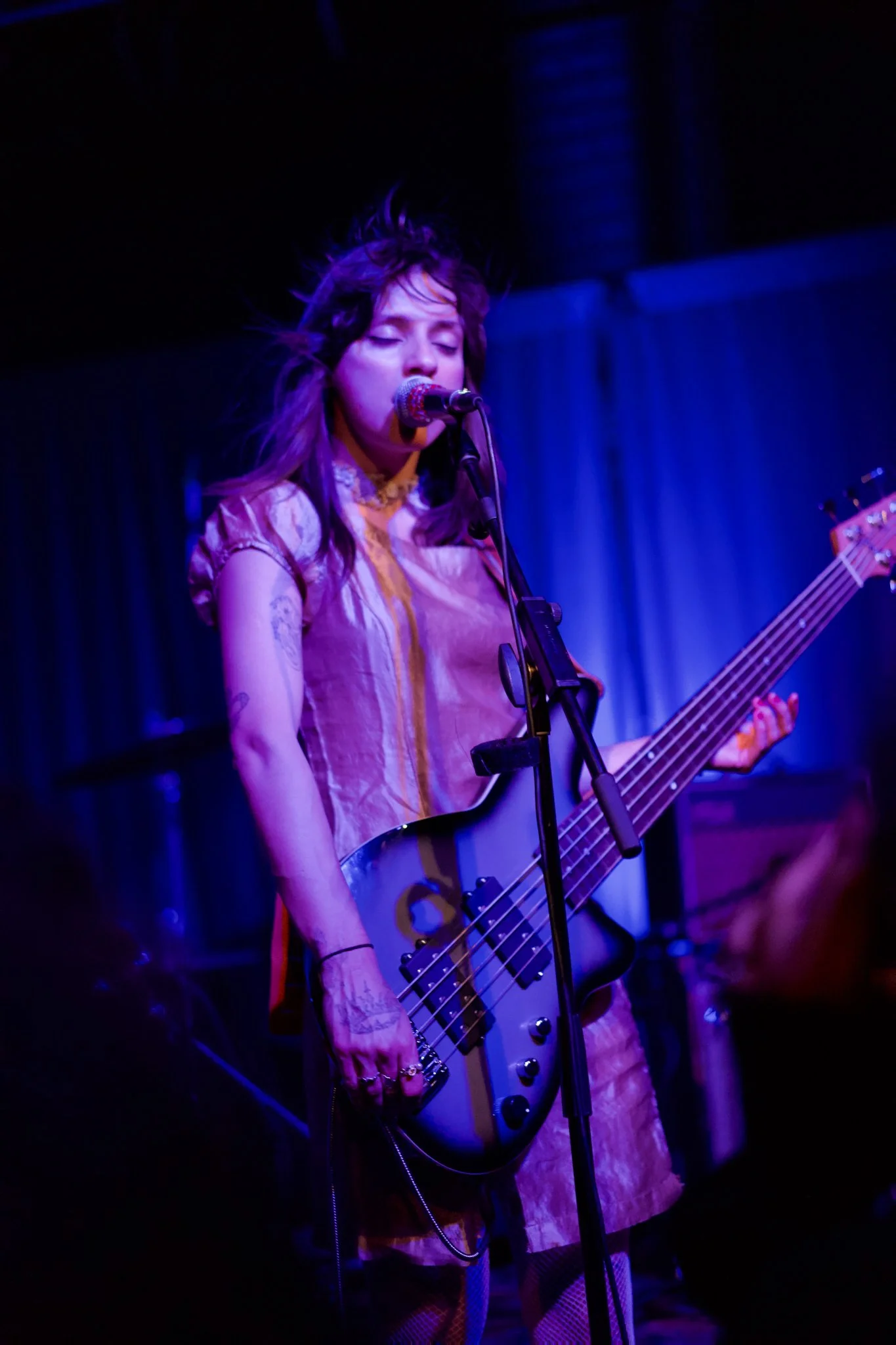A female musician with purple hair sings into a microphone while playing a bass guitar on stage at a dimly lit venue.