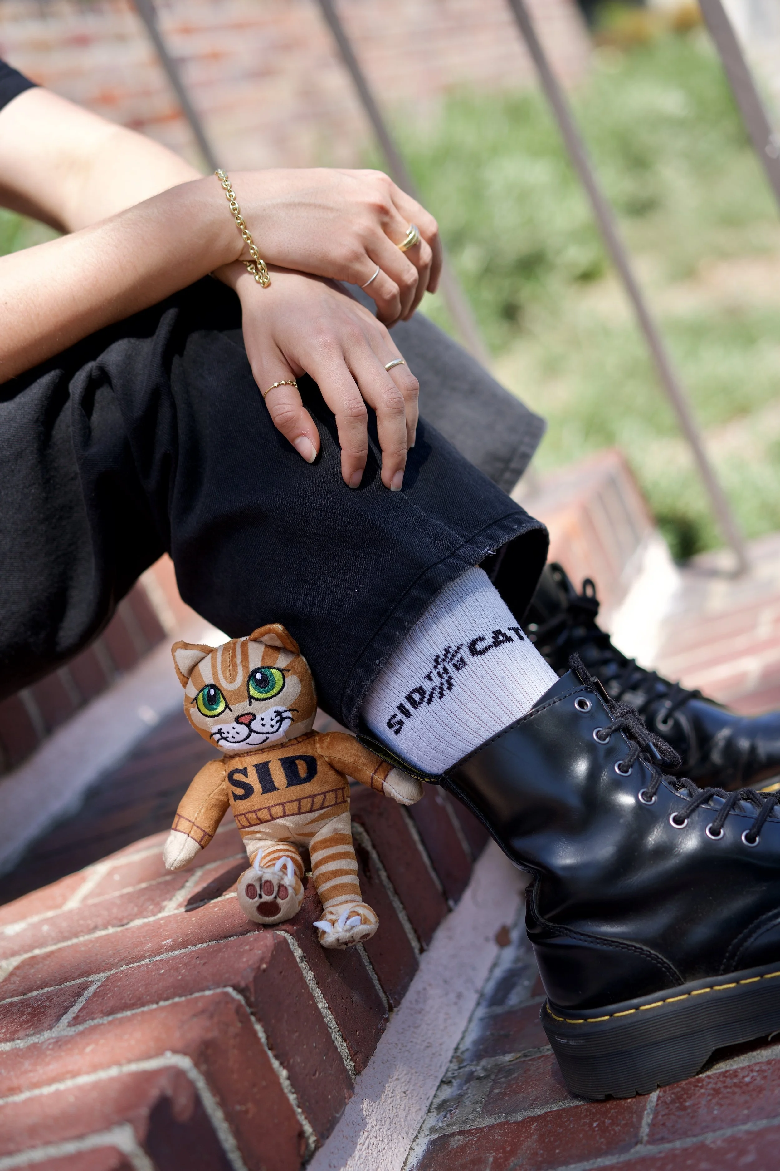 Close-up of a person's legs and hands resting on their lap, wearing black boots, black shorts, white socks with black text, and gold rings, sitting on a brick ledge with a green and red brick background. A plush cat toy with orange and white stripes, green eyes, and “SID” written on its body is leaning against the brick ledge.