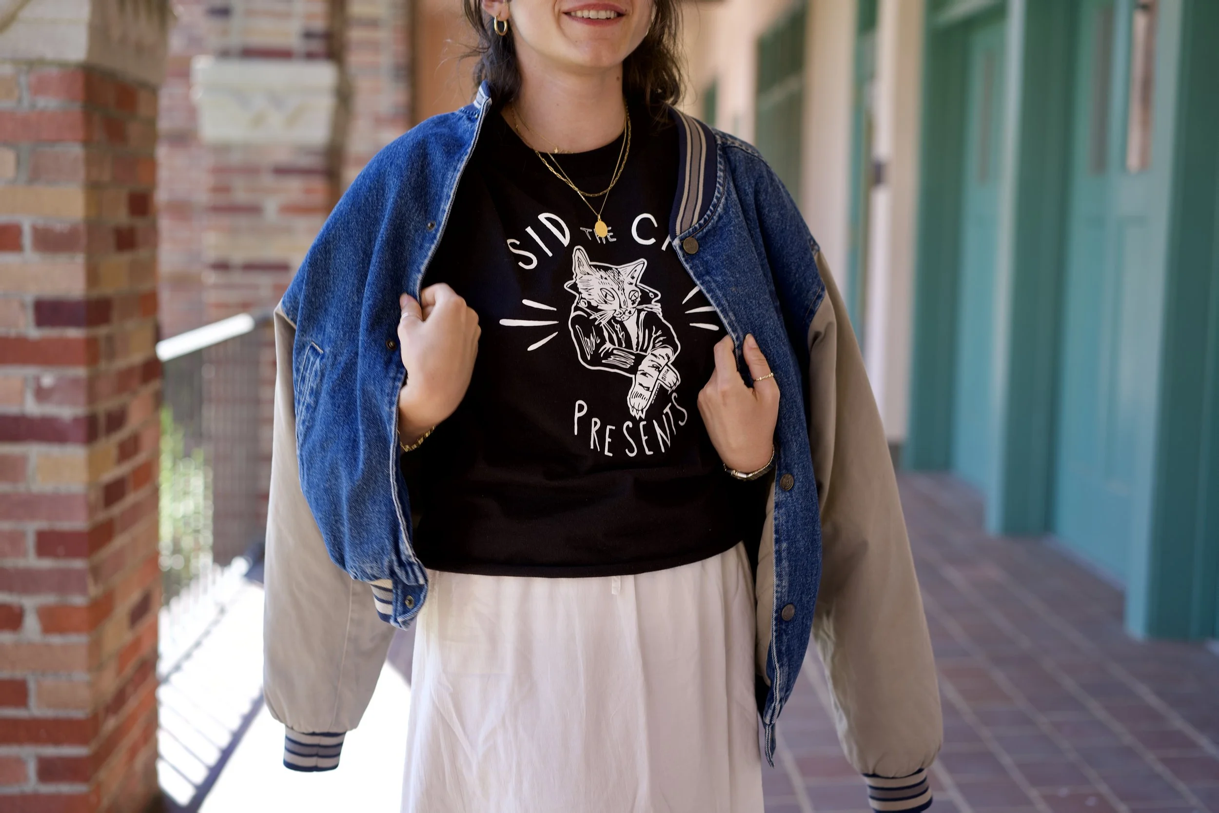 A person smiling, wearing a black shirt with a graphic of a cat and the text "Sidi C Presents," a denim jacket, beige skirt, striped socks, layered necklaces, and gold hoop earrings, standing outside near a brick pillar and green lockers.