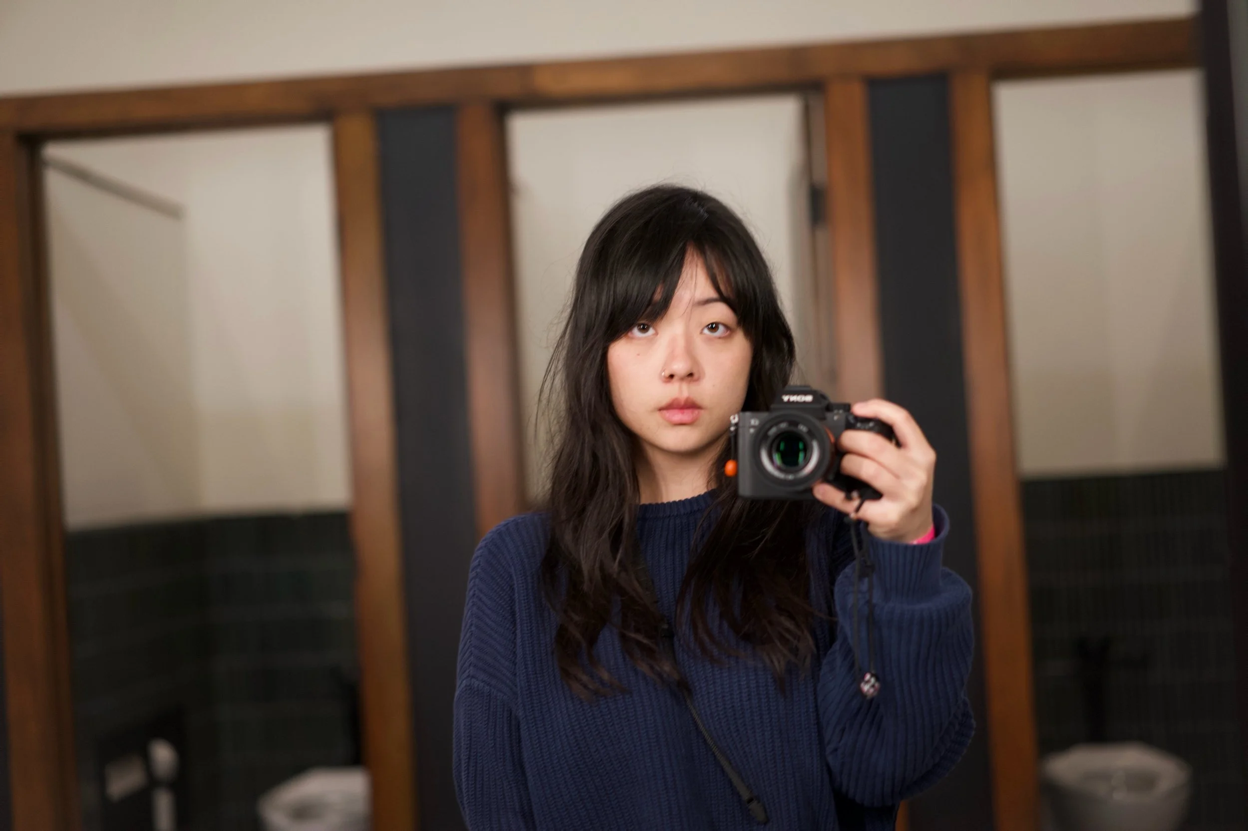 Young woman with black hair taking a selfie in a mirror, holding a camera, standing in a room with wooden beams