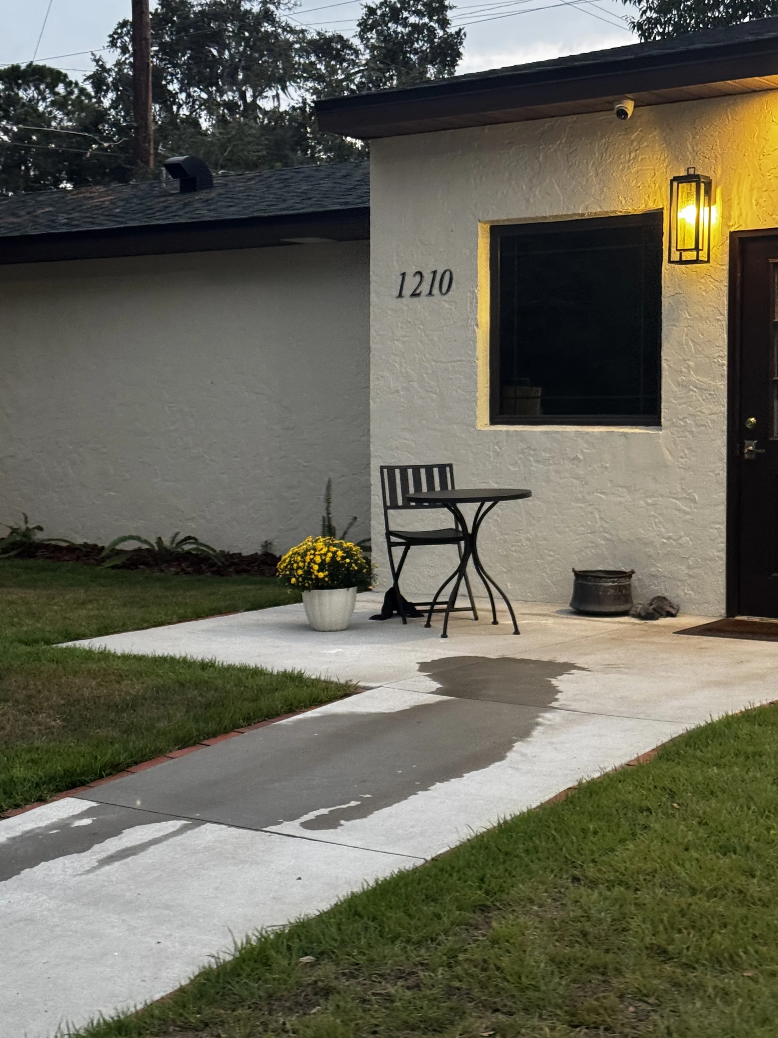A house with the number 1210 on the wall, front porch with a small table and chair, yellow outdoor light, potted yellow flowers, and a wet sidewalk surface at dusk.