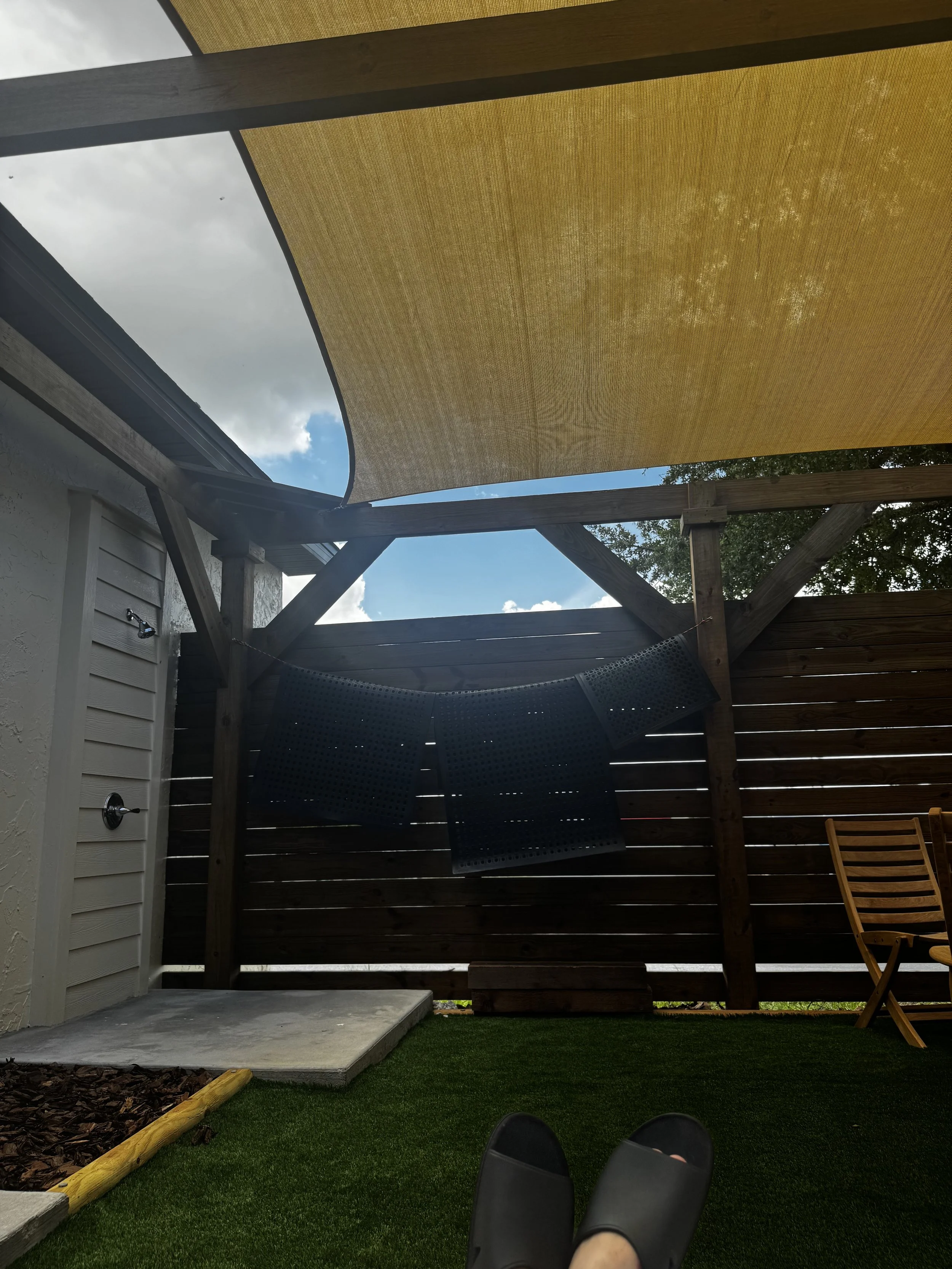 Person's feet in black slides viewed from a backyard patio with artificial grass, a wooden privacy fence, a yellow shade sail, and a cloudy sky.