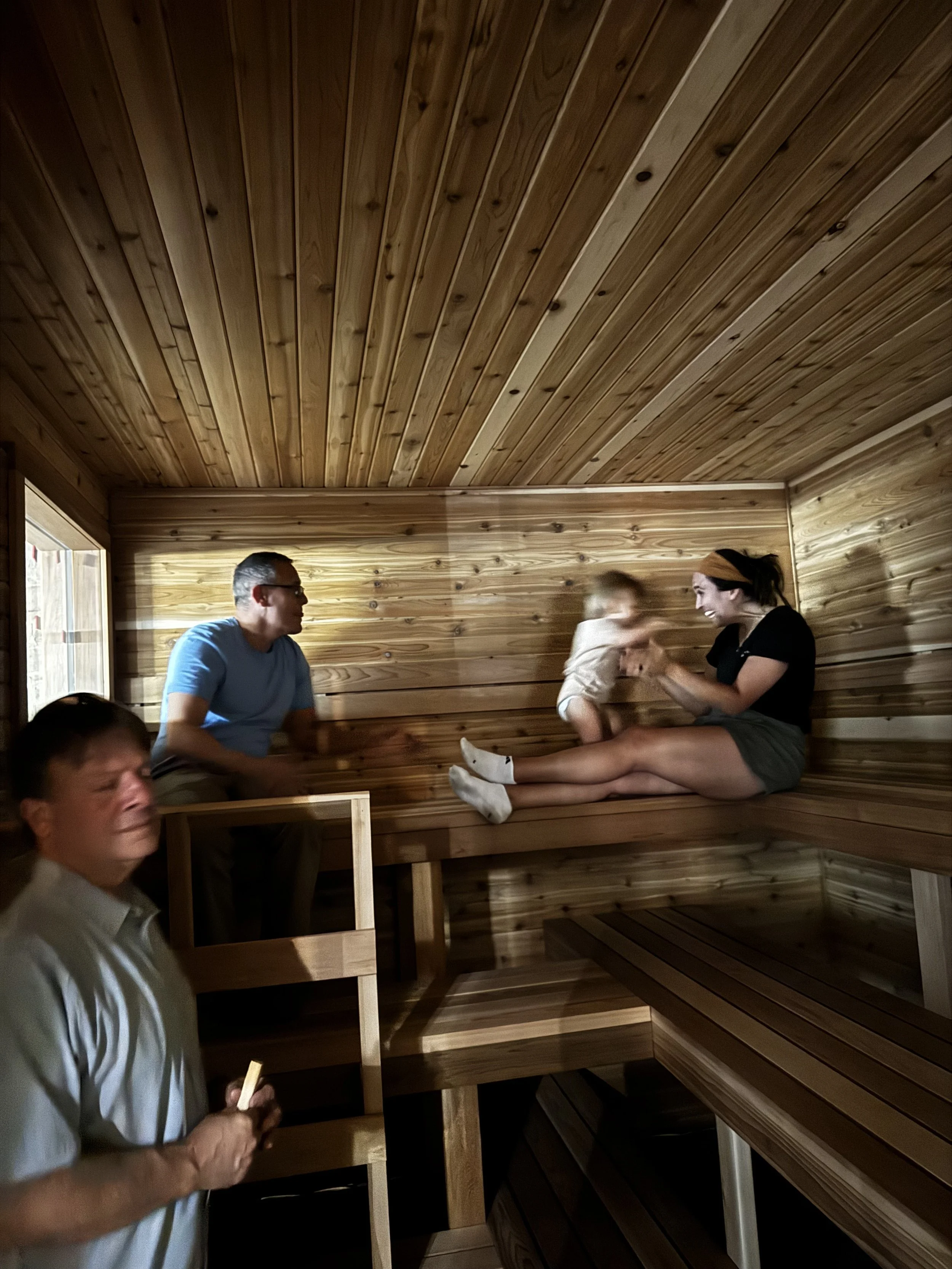 People relaxing and chatting inside a wooden sauna, with a man standing and holding a wooden ladle or handle, and a woman sitting on a bench smiling and engaging in conversation.