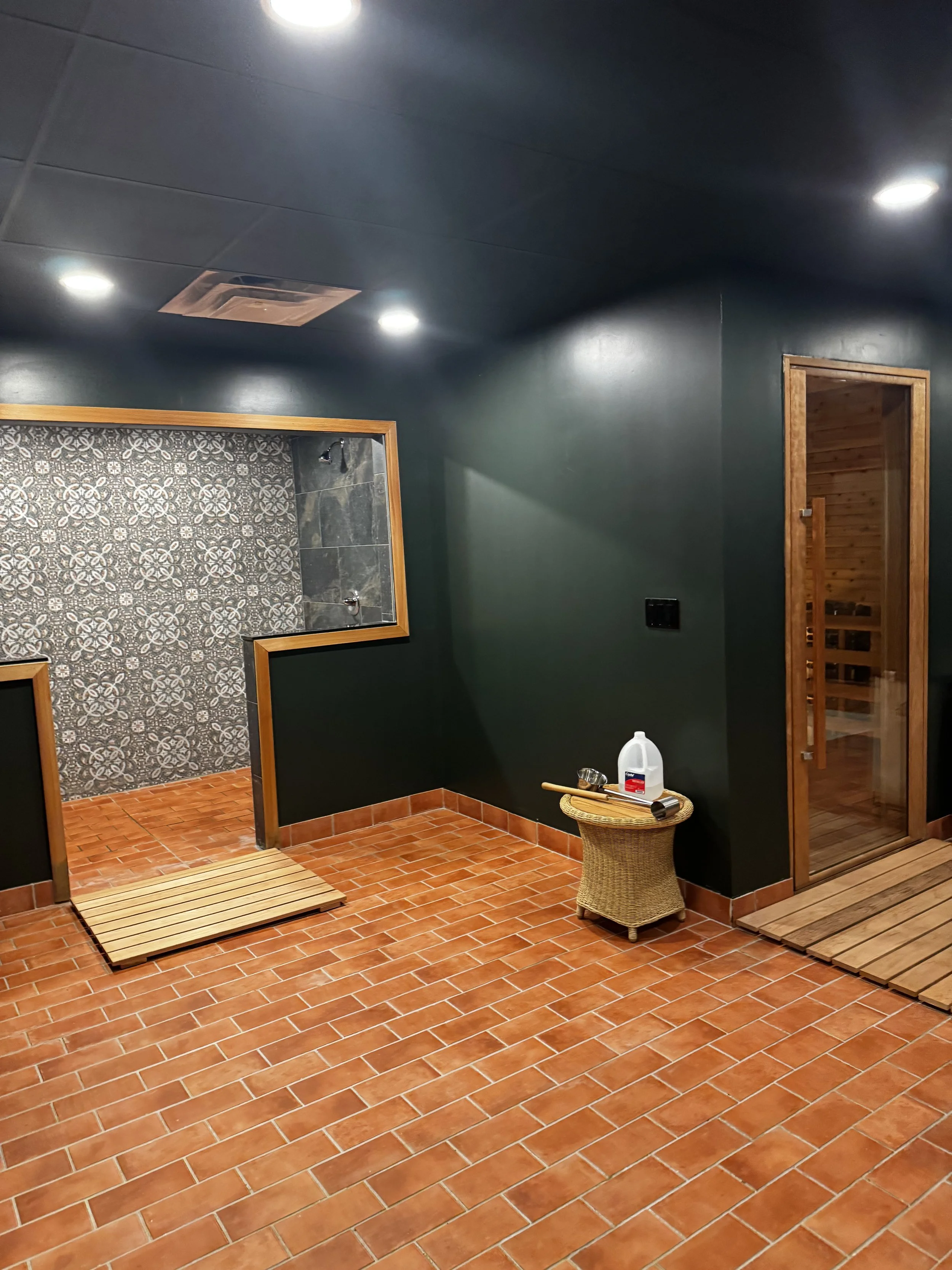 A room with dark green walls and a reddish-brown tiled floor, featuring a small shower area with patterned tiles, a wooden-framed doorway leading to a sauna, and a wicker table with a white water container and a metal scoop.