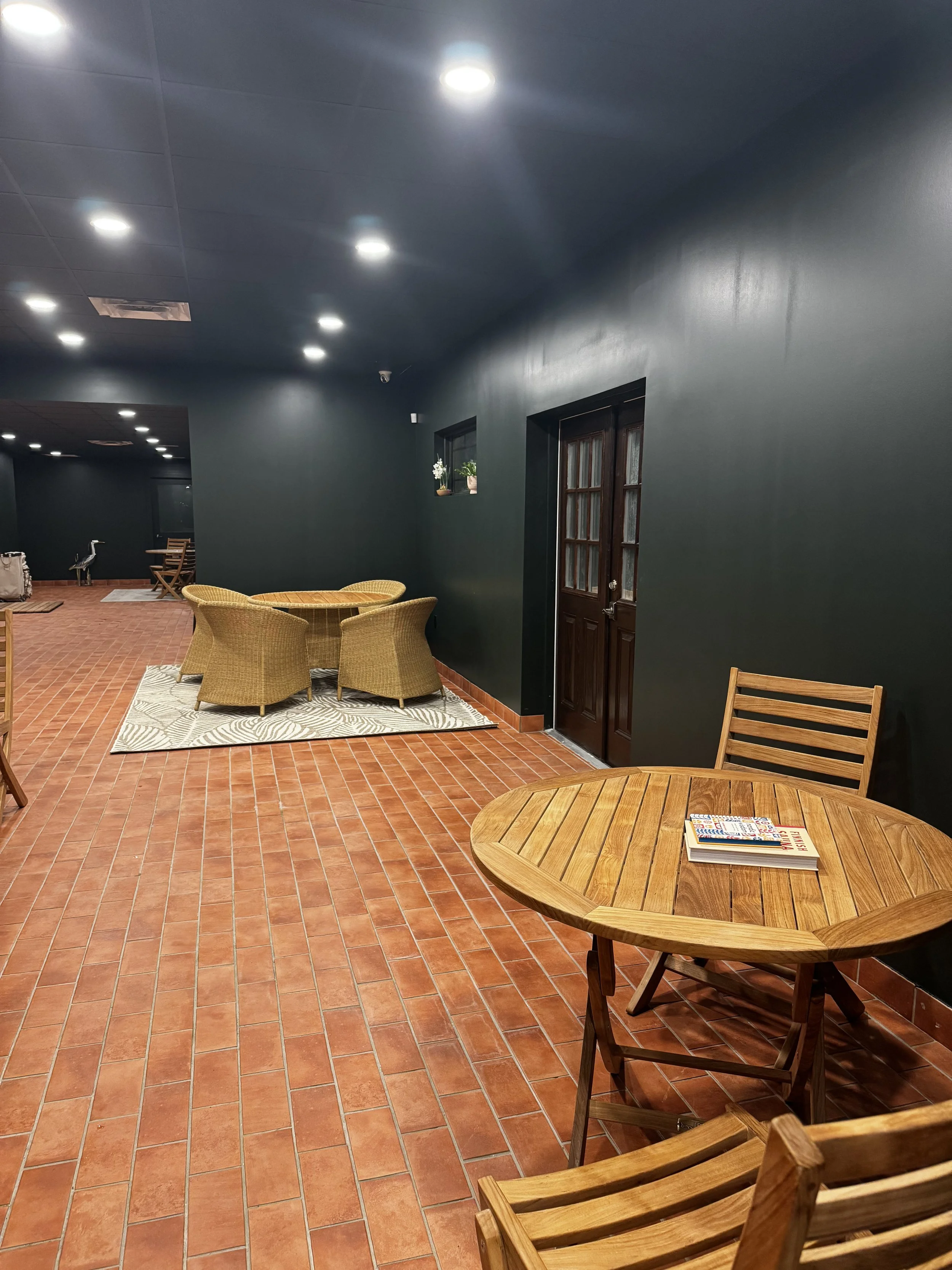 Indoor seating area with red tile flooring, a round wooden table with two wooden chairs, another table with wicker chairs, and windows with potted plants, dark painted walls, and ceiling lights.