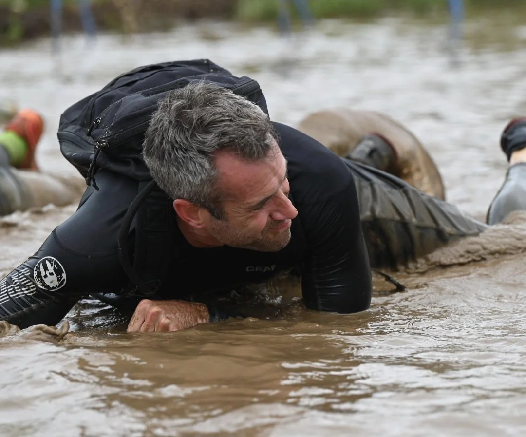Health coach Brett Thomas crawling under barbed wire with a GoRuck bag during an OCR, demonstrating functional movement and physical resilience.