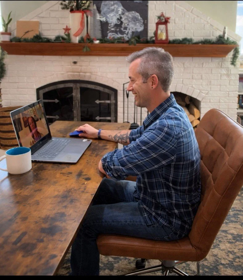 Brett Thomas smiles during a video call at a wooden desk in a professional home office setting, with a laptop, coffee mug, and white brick fireplace in the background.