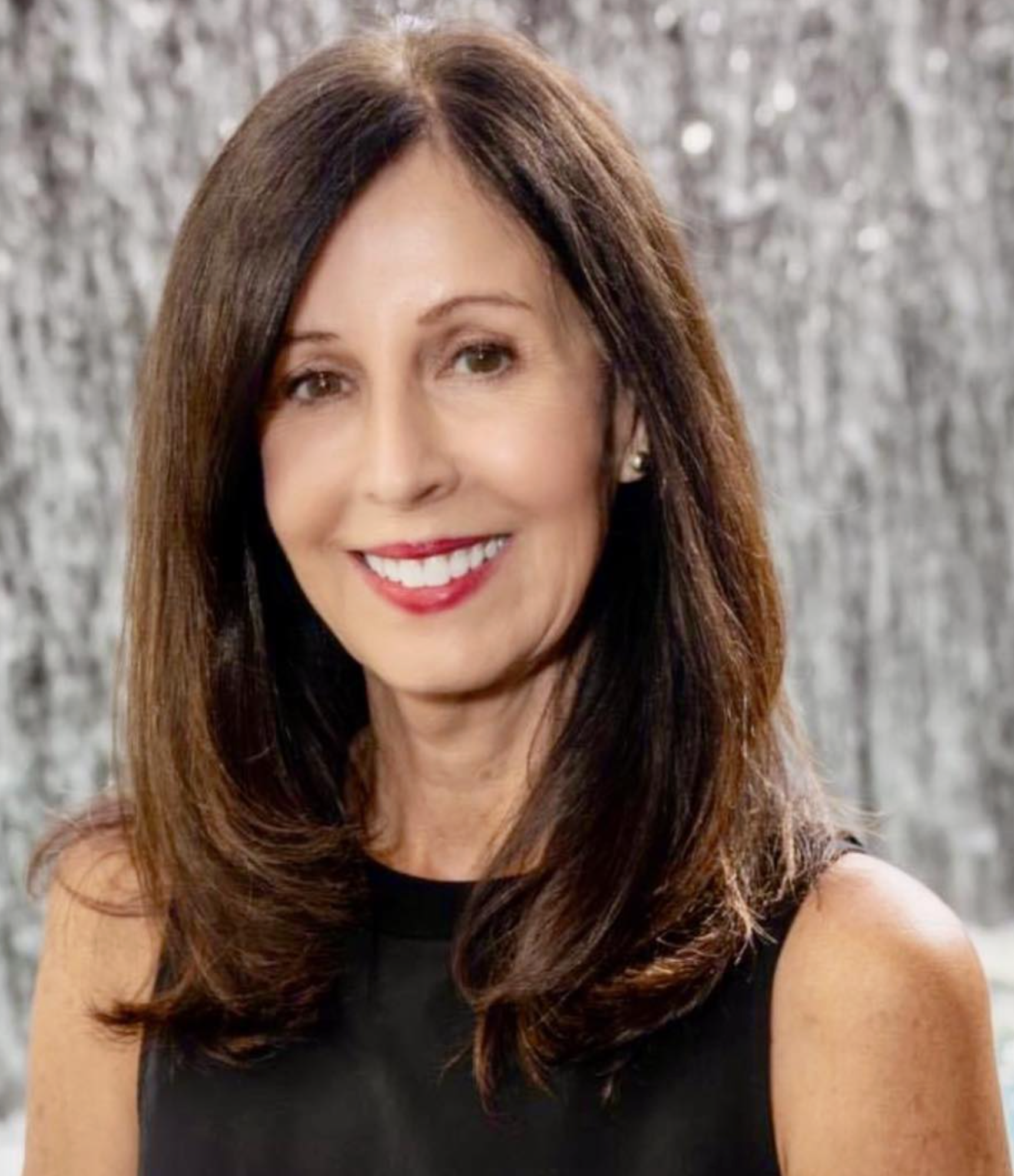 Joni Johnston Neidagh - A woman with shoulder-length brown hair, wearing a sleeveless black top, smiling in front of a silver glittery background.