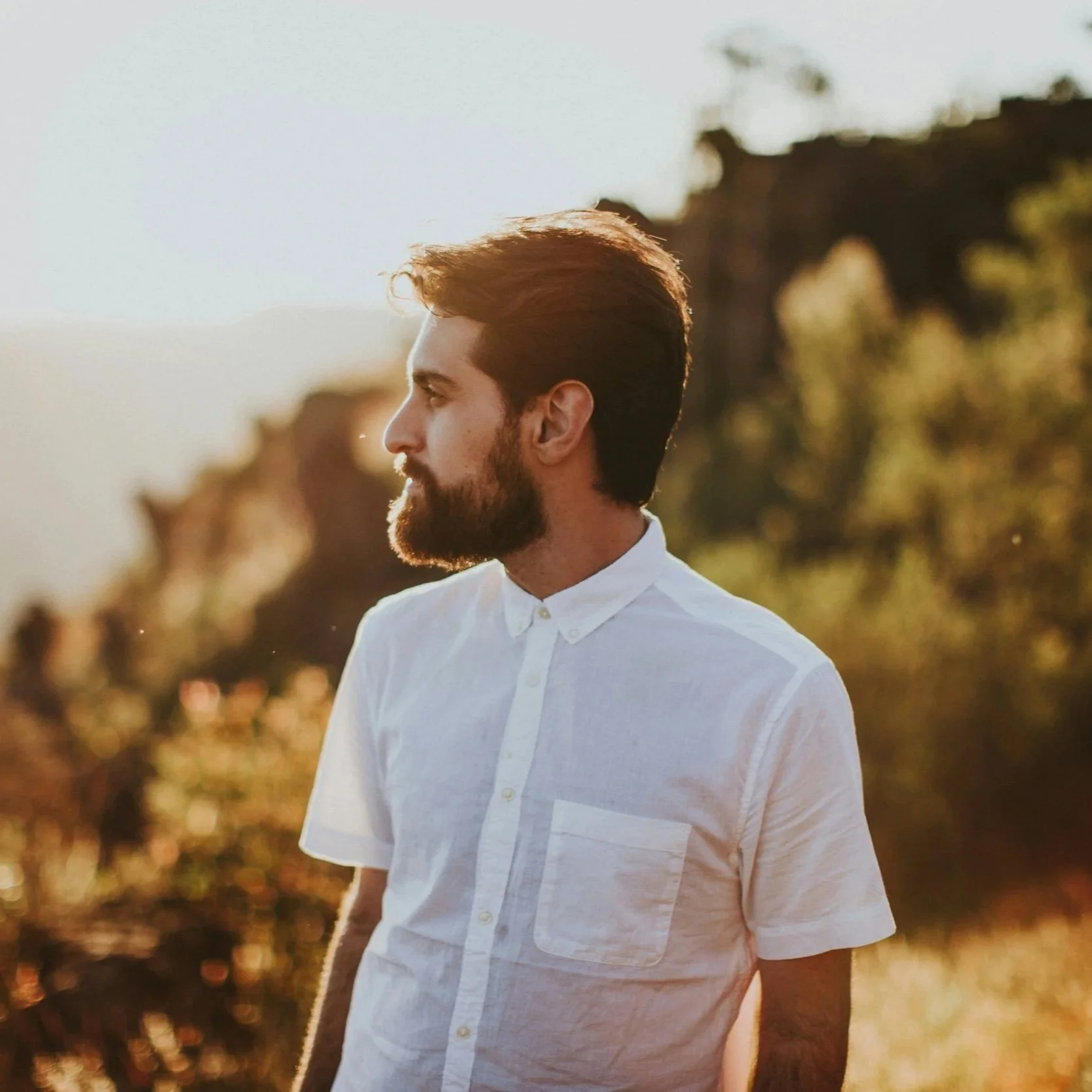 A man with brown hair and a beard wearing a white button-up shirt, looking to the left, outdoors during sunset with trees and a bridge in the background.
