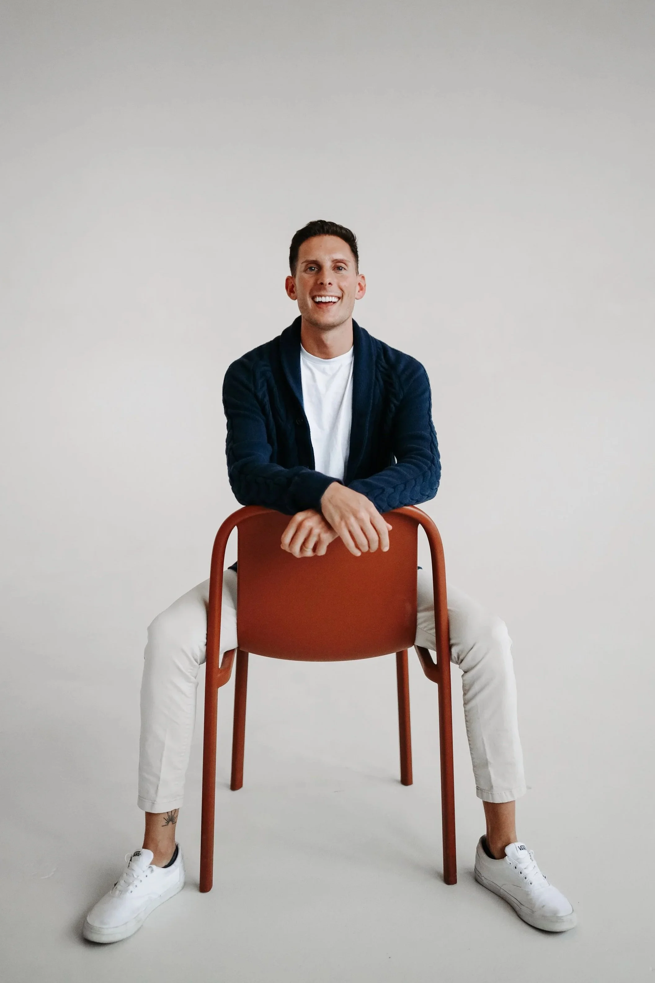 A man sitting on a chair with legs spread apart and arms resting on the back of the chair, smiling, wearing a navy blue cardigan, white T-shirt, white pants, and white sneakers, with a small tattoo on his left ankle, against a plain white background.