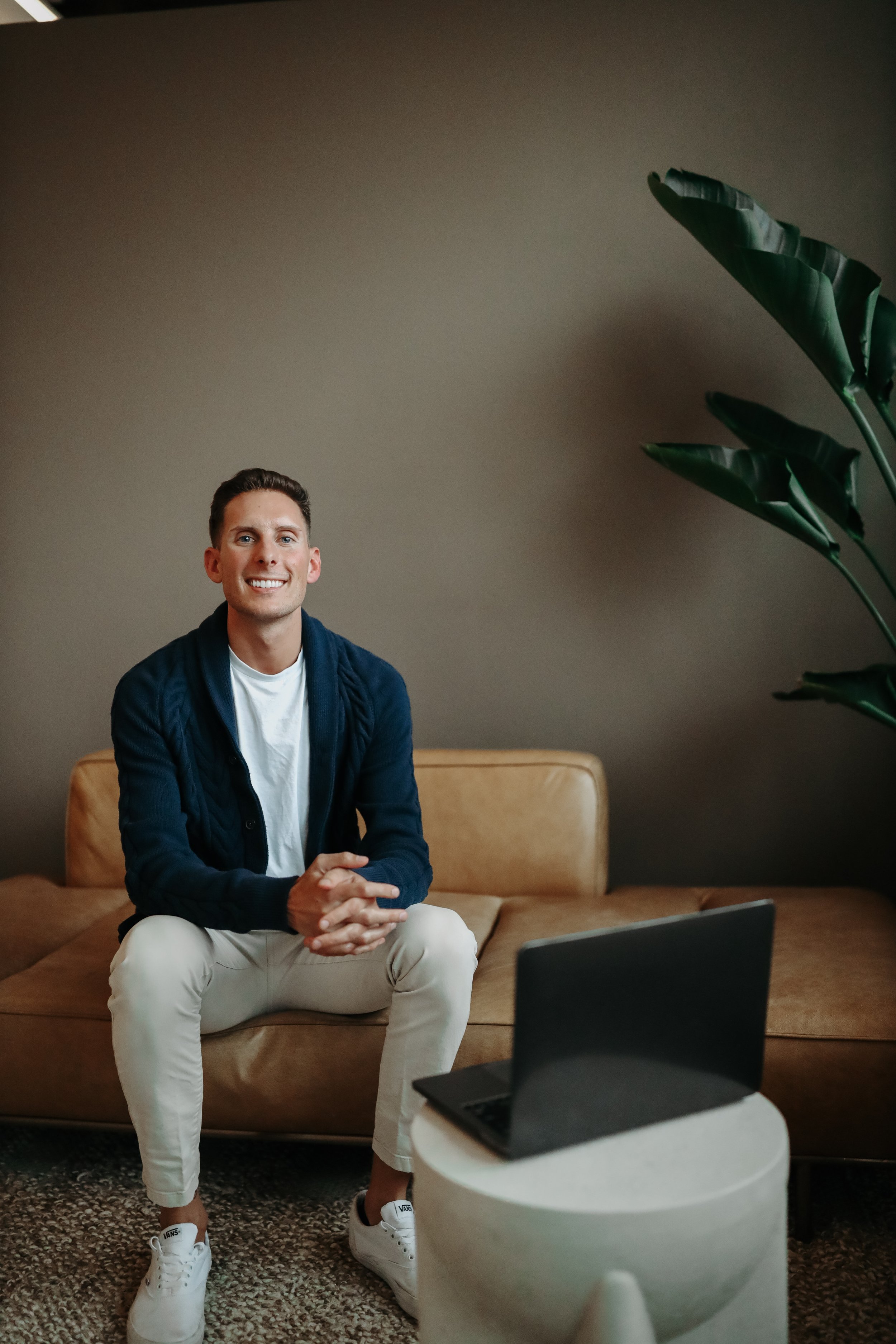 A young man with short brown hair and light skin sitting on a tan couch in front of a beige wall. He is smiling, wearing a white t-shirt, a navy blue cardigan, and white sneakers. There is a large green plant to his right and a laptop on a small round table in front of him.