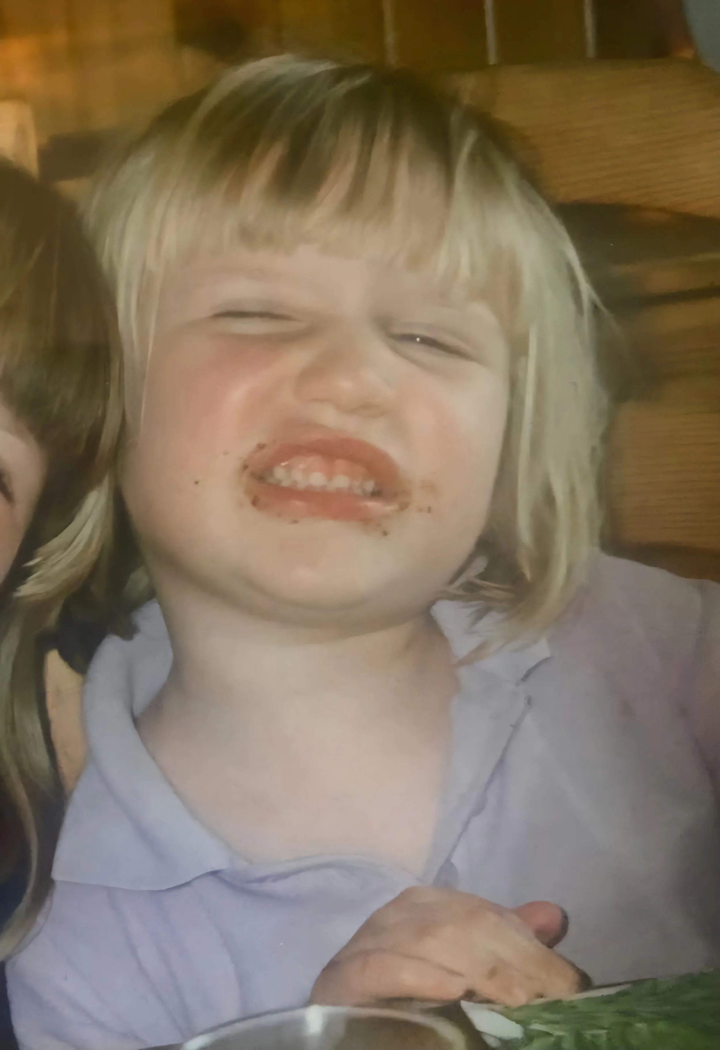 A young child with blond hair and a big smile, lying on a white surface, with part of a person's arm or leg visible at the bottom, and a wooden surface in the background.