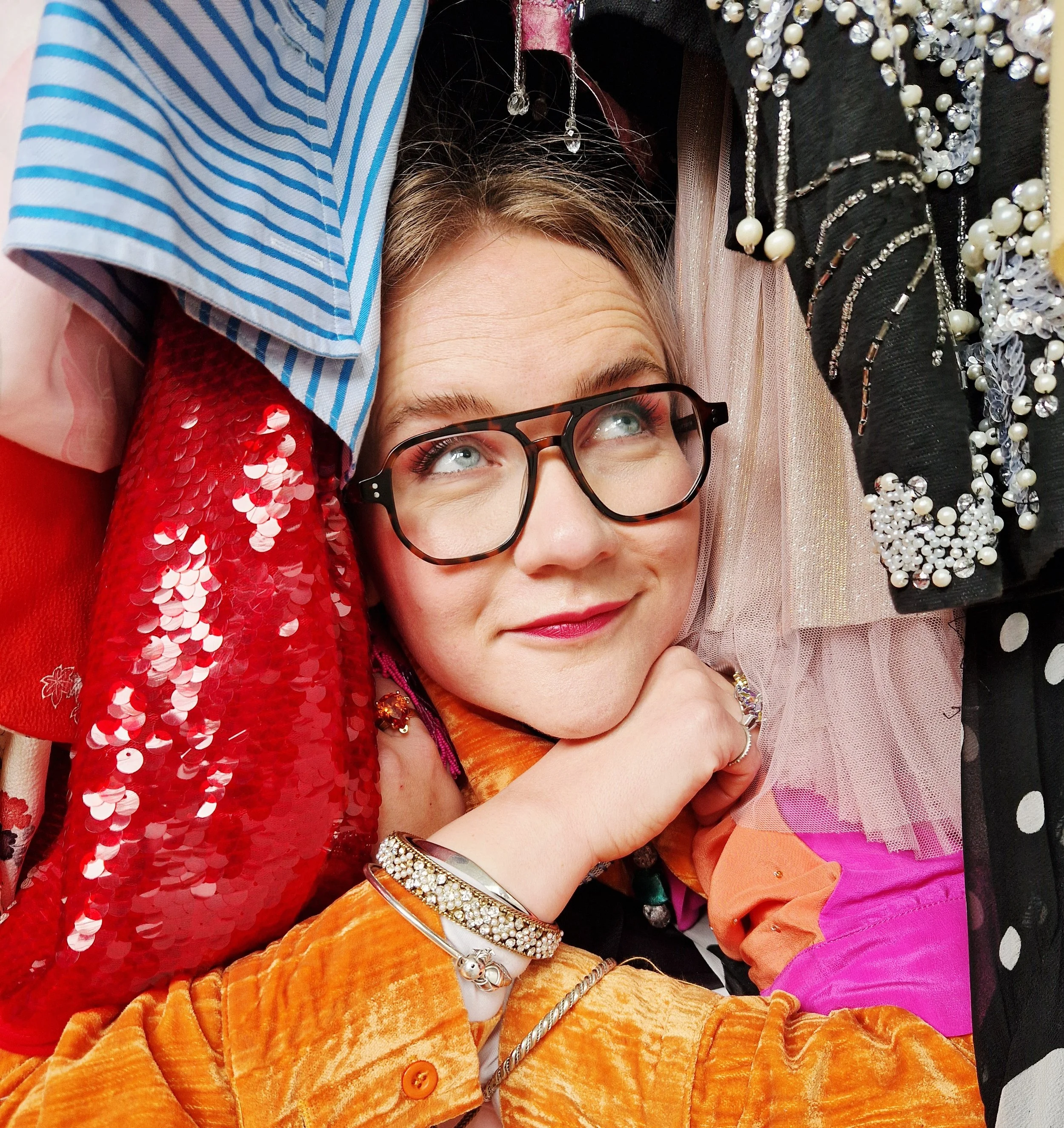 Close-up of a woman with glasses and earrings, lying on a colorful assortment of fabrics and accessories, including sequined, pearl, and beaded embellishments, with vibrant colors and textures.