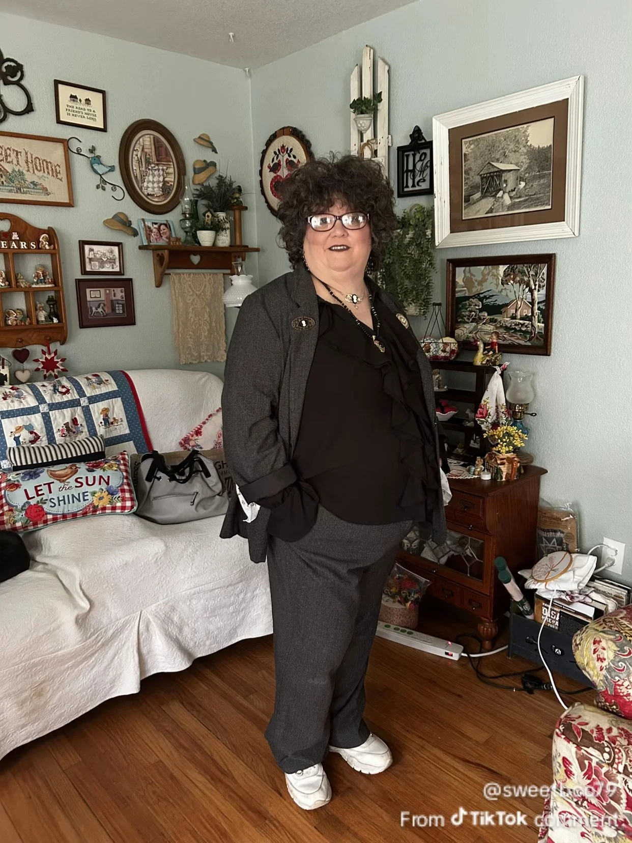 A woman with curly hair and glasses standing in a living room with decorated walls including framed pictures, a quilt, and various knickknacks on shelves and furniture.