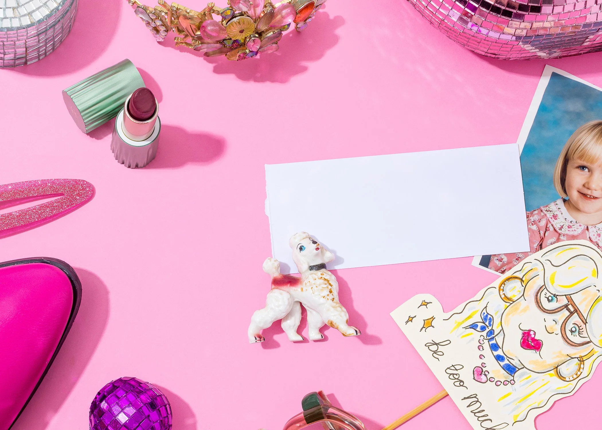 A flat lay of various items on a pink background, including a lipstick in a pastel pink and green case, a pink glittery hair clip, purple disco ball ornaments, a photograph of a smiling girl with blonde hair, a white paper with doodles, a small unicorn figurine, and decorative jewelry pieces.