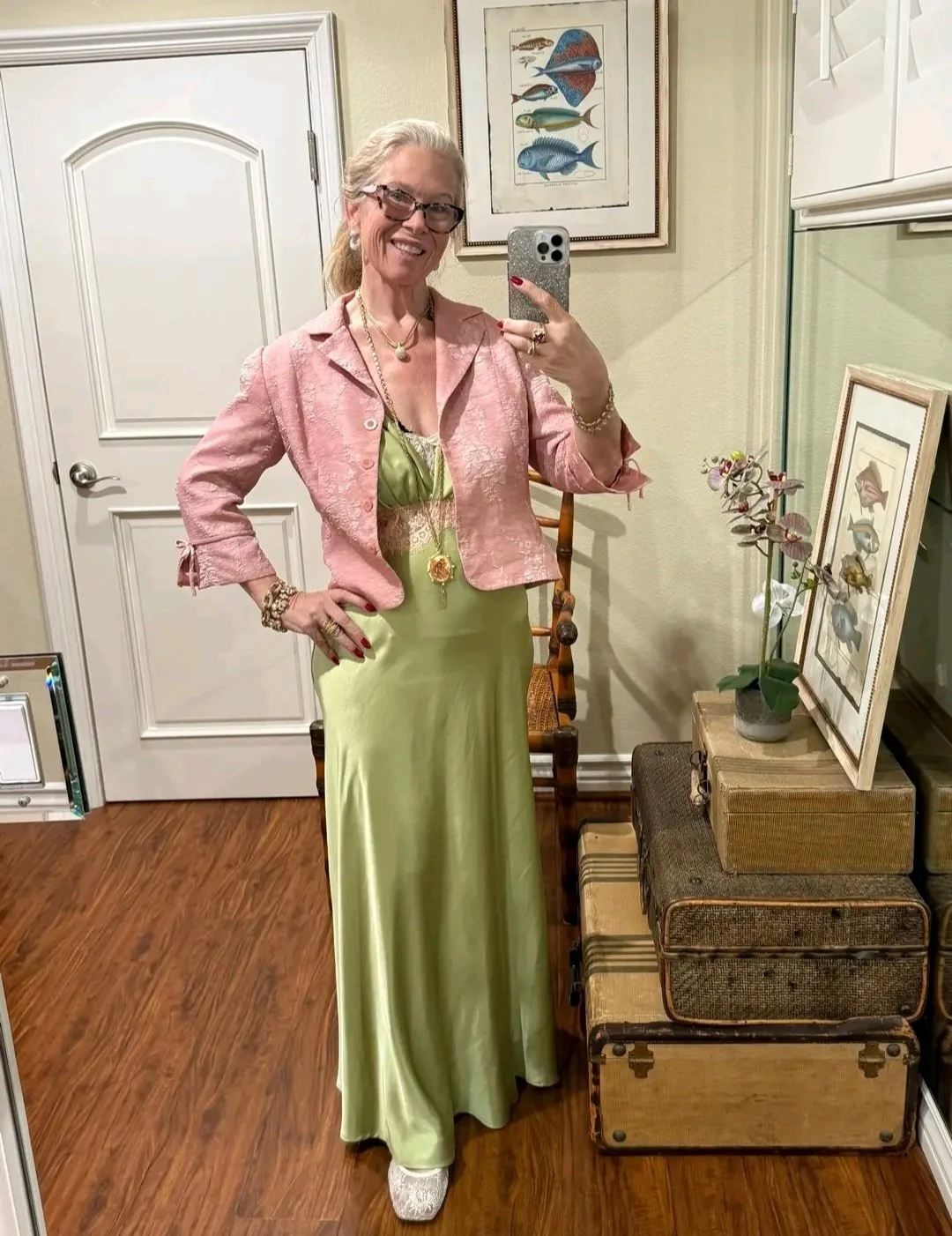 A woman taking a selfie in a mirror, wearing a pink jacket over a green dress, with jewelry, glasses, and smiling.