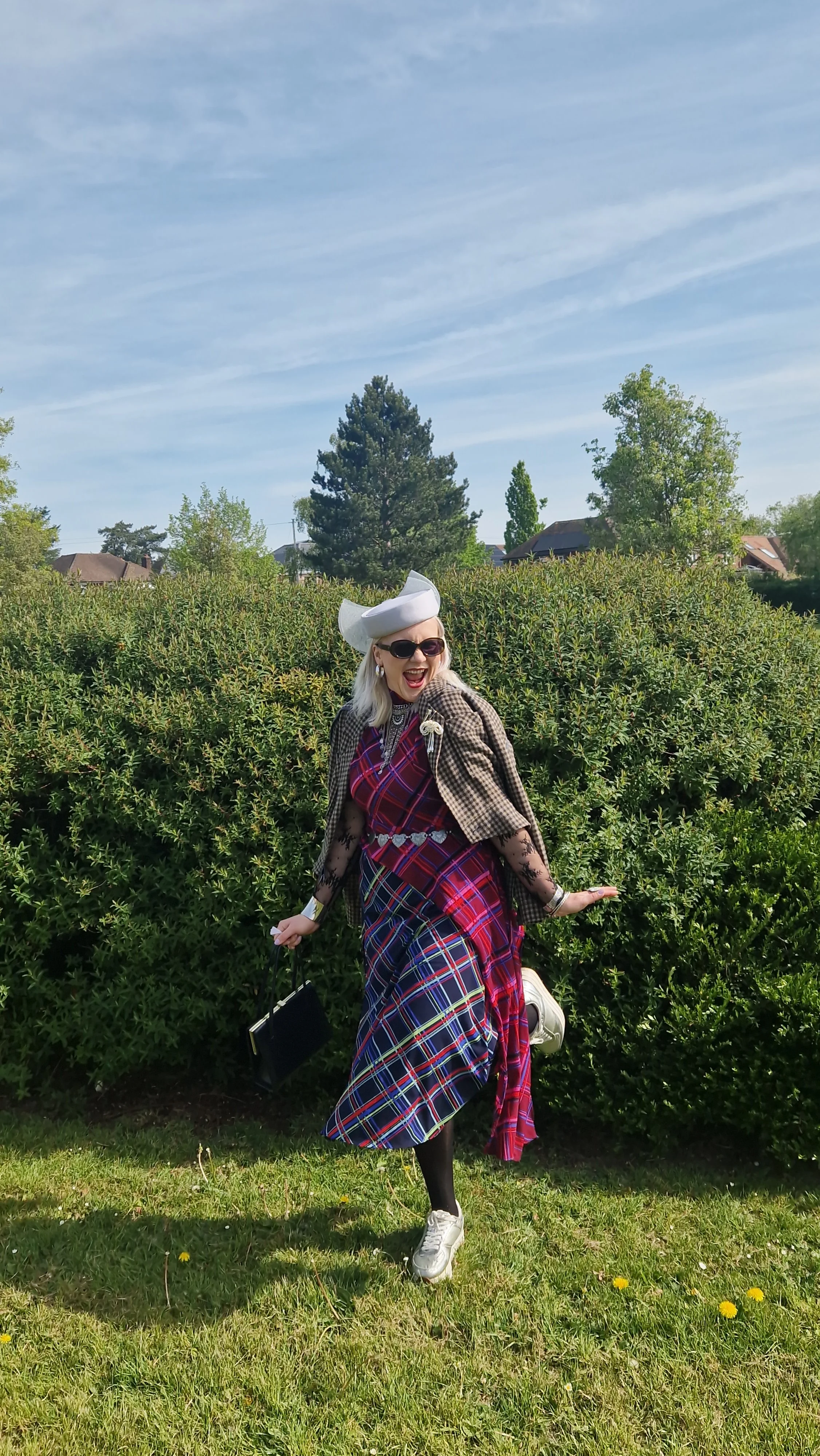 A woman in colorful tartan clothing, wearing a white hat, sunglasses, and black lace gloves, standing outdoors in front of greenery, smiling and striking a playful pose.