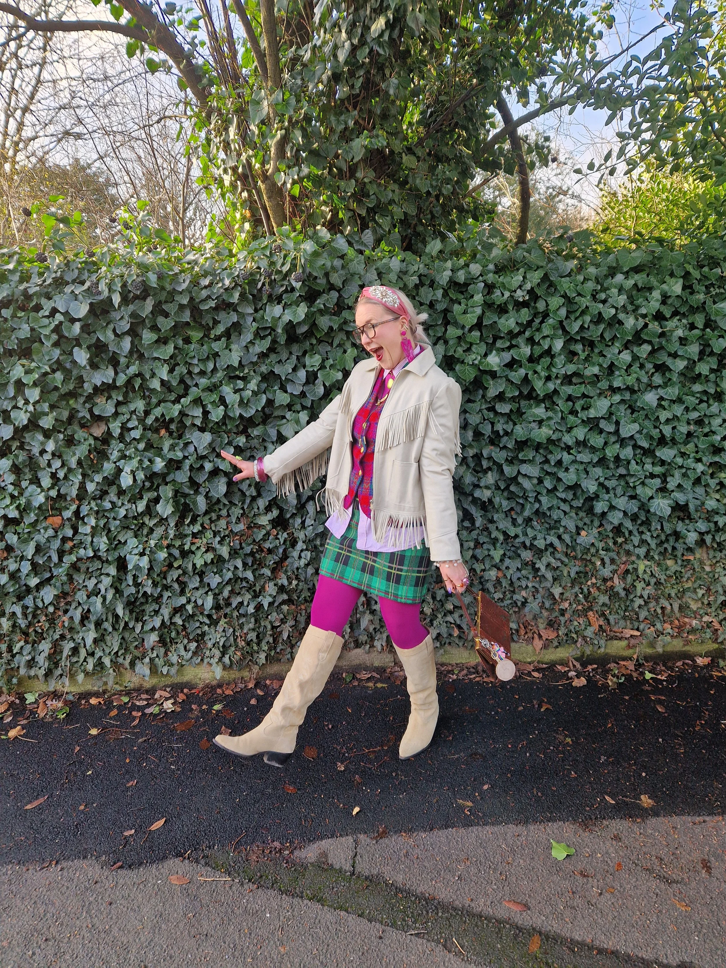 Woman in beige fringed jacket and beige boots, wearing pink tights, a colorful plaid skirt, and earrings, standing next to a hedge with a tree in the background. She is smiling and looking down, holding a small handbag.