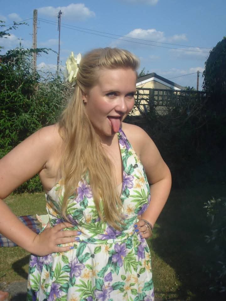 Woman with long blonde hair wearing a floral dress with purple, green, and yellow flowers, sticking her tongue out outdoors on a sunny day.