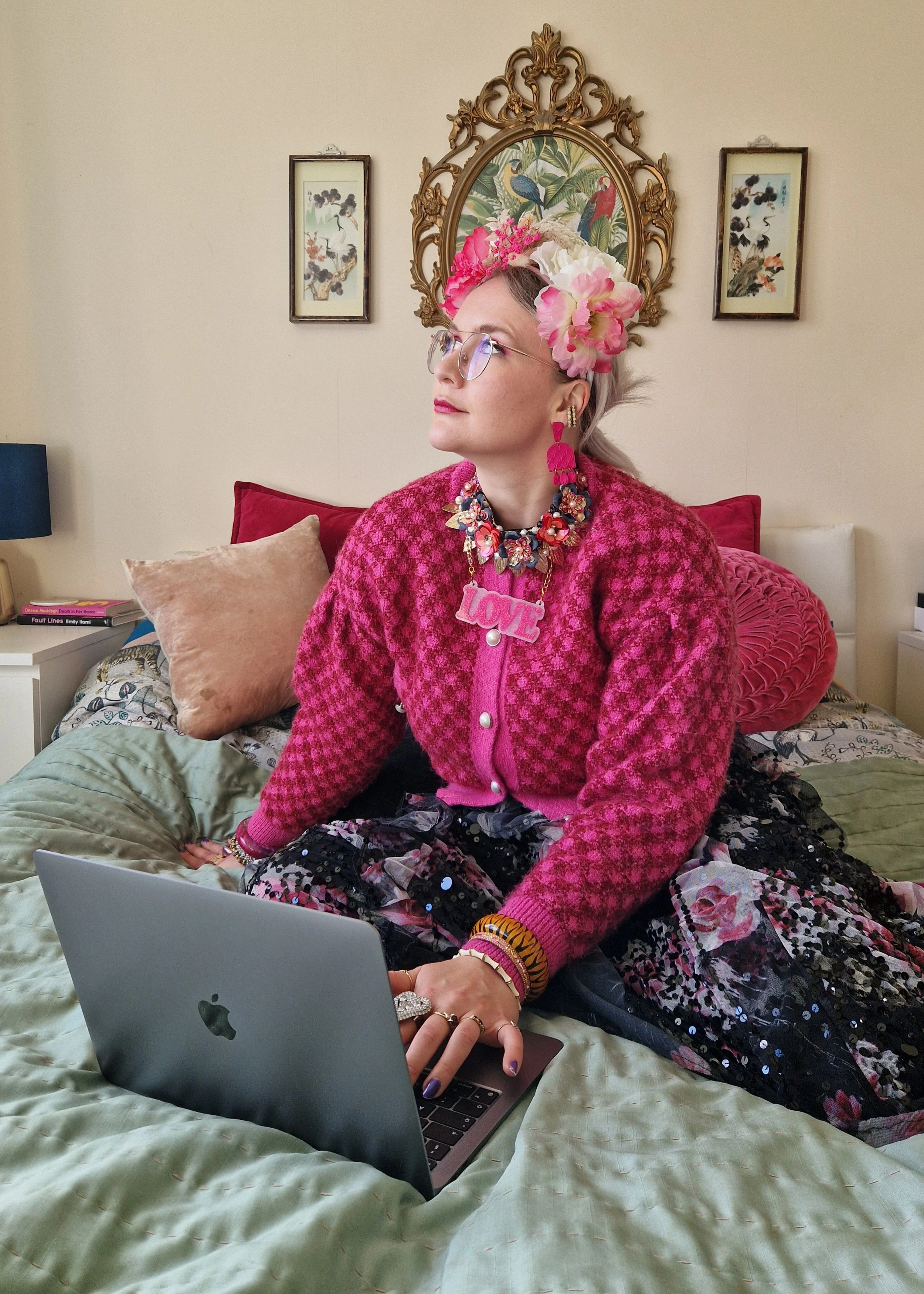 A woman sitting on a bed, working on a laptop. She has light gray hair, glasses, and wears a pink textured jacket, jewelry, and a floral headband. The room has beige walls, framed art, and pillows on the bed.