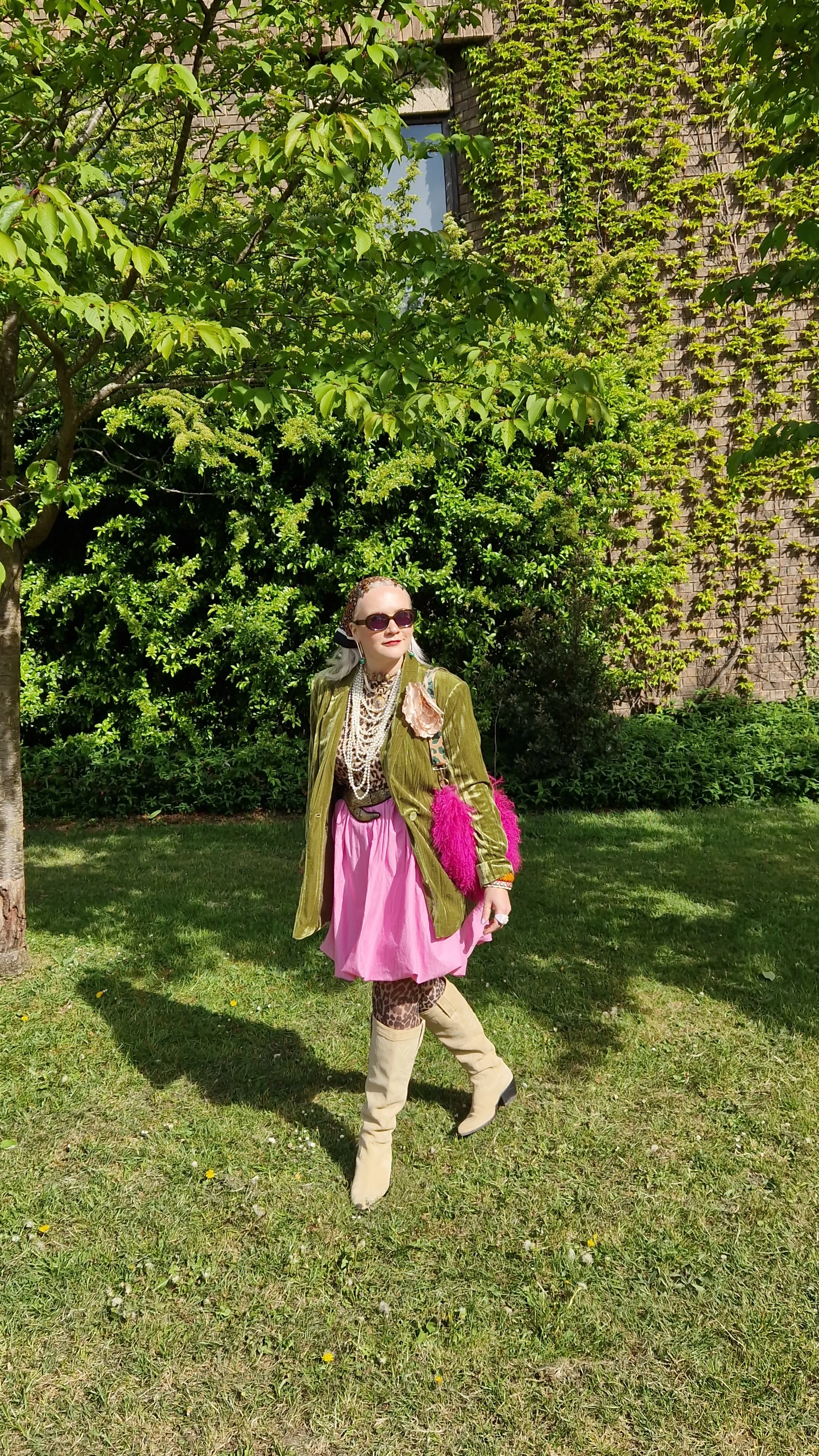 A woman standing outdoors in a garden area with green trees and a brick building, wearing sunglasses, a green jacket, a pink skirt, leopard print tights, beige boots, and accessorized with jewelry and a pink furry bag.