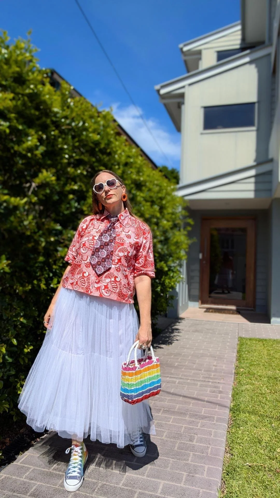 A woman standing outside in front of a house, wearing pink sunglasses, a red patterned shirt with a tie, a white tulle skirt, colorful sneakers, and holding a rainbow striped bag.