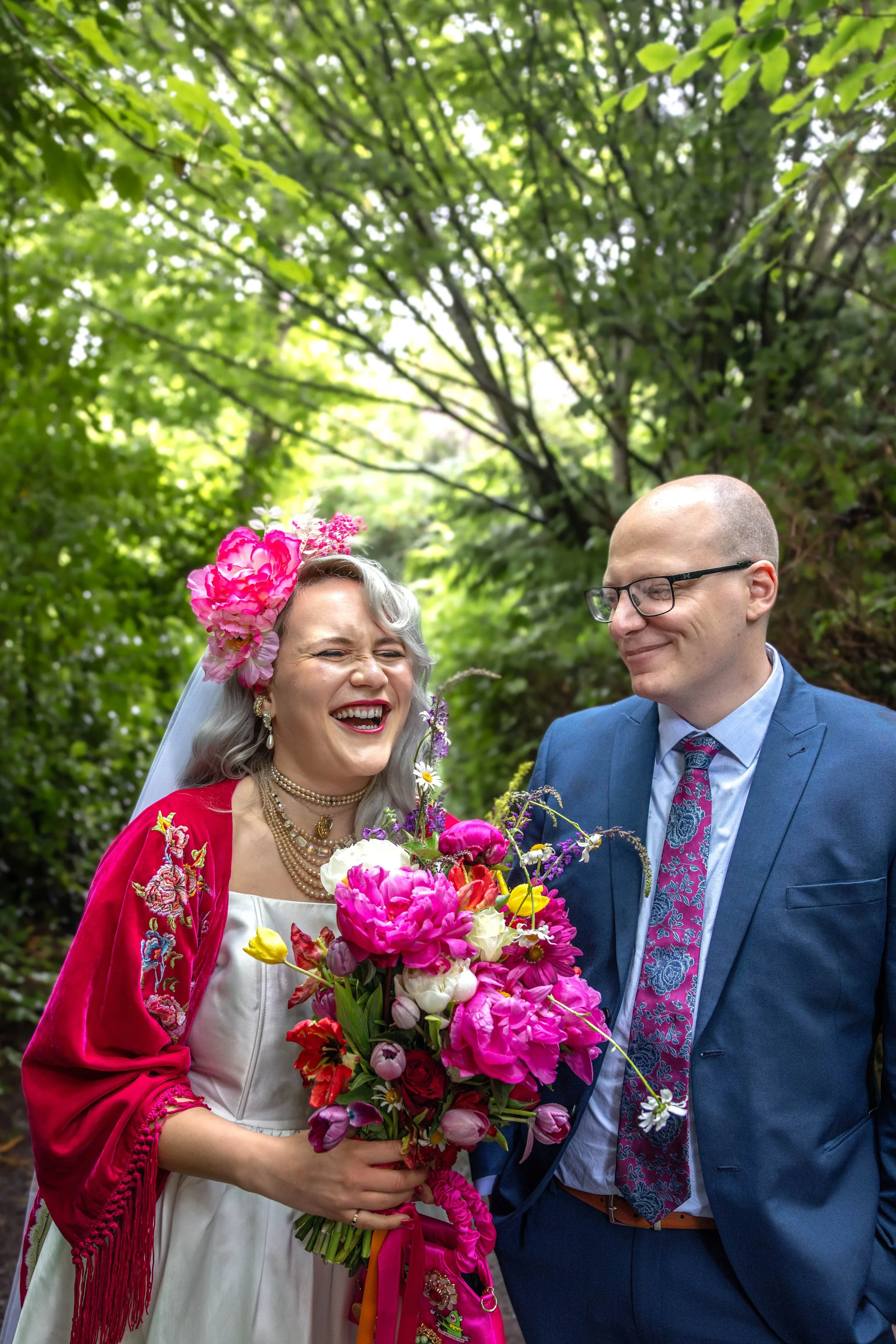 A couple dressed in colorful wedding attire, holding a large bouquet of vibrant pink, red, yellow, and white flowers, standing outdoors in a lush green wooded area during daytime.