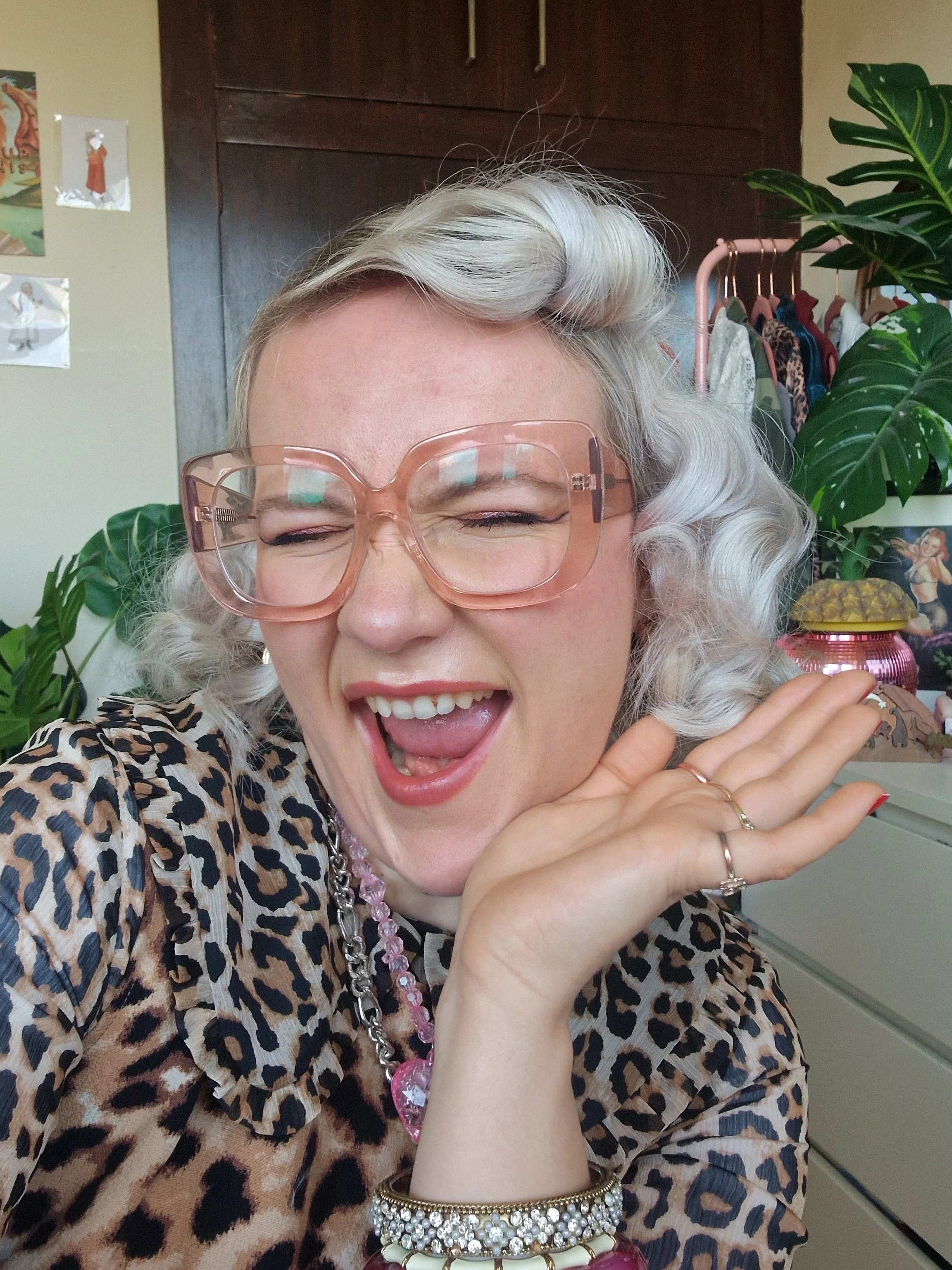 A woman with light blonde hair and glasses smiling and posing in a room with plants and clothing rack in the background.