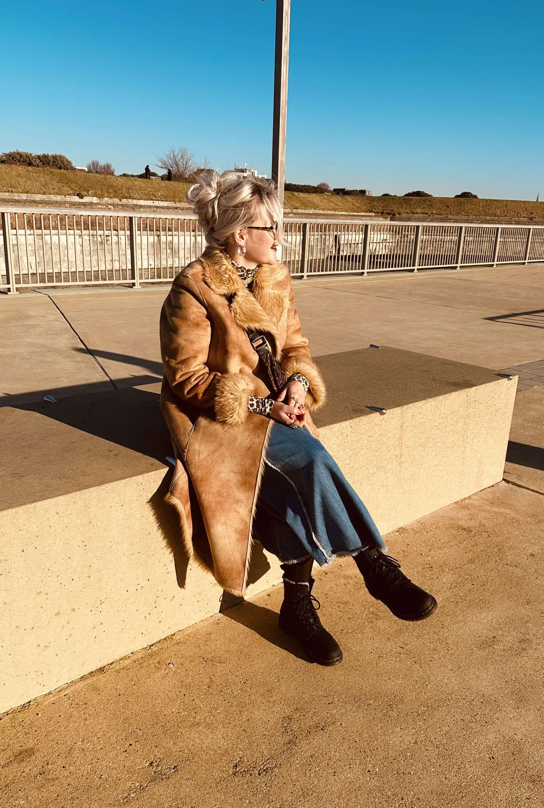 A woman with  hair in a messy bun, wearing glasses, earrings, a leopard print turtleneck, a long brown fur-lined coat, jeans, and black boots, sitting on a concrete bench outdoors on a sunny day, with a clear blue sky and grassy hill in the background.