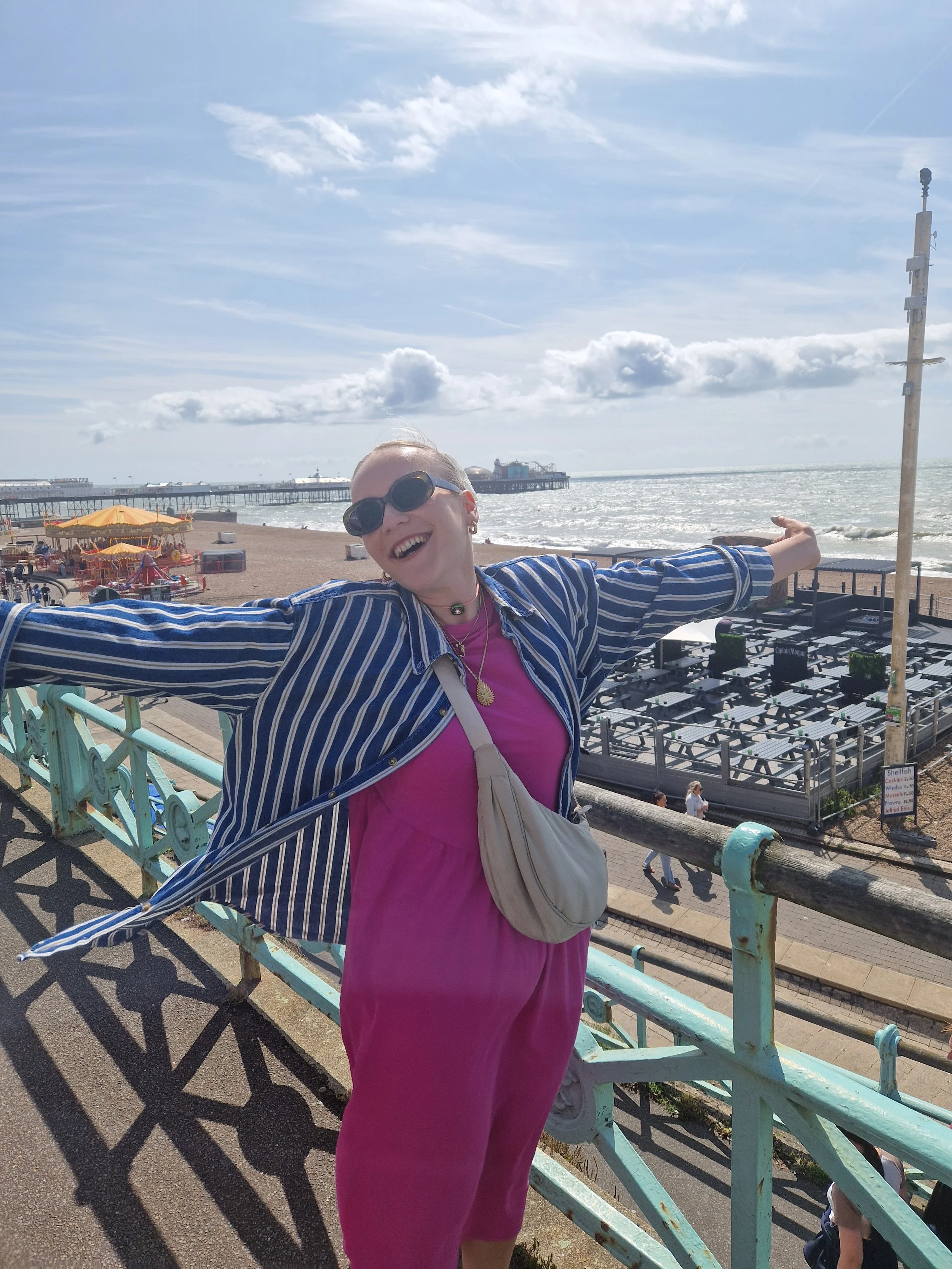 A woman with glasses and a striped jacket smiling with her arms outstretched on a bridge over a beach.