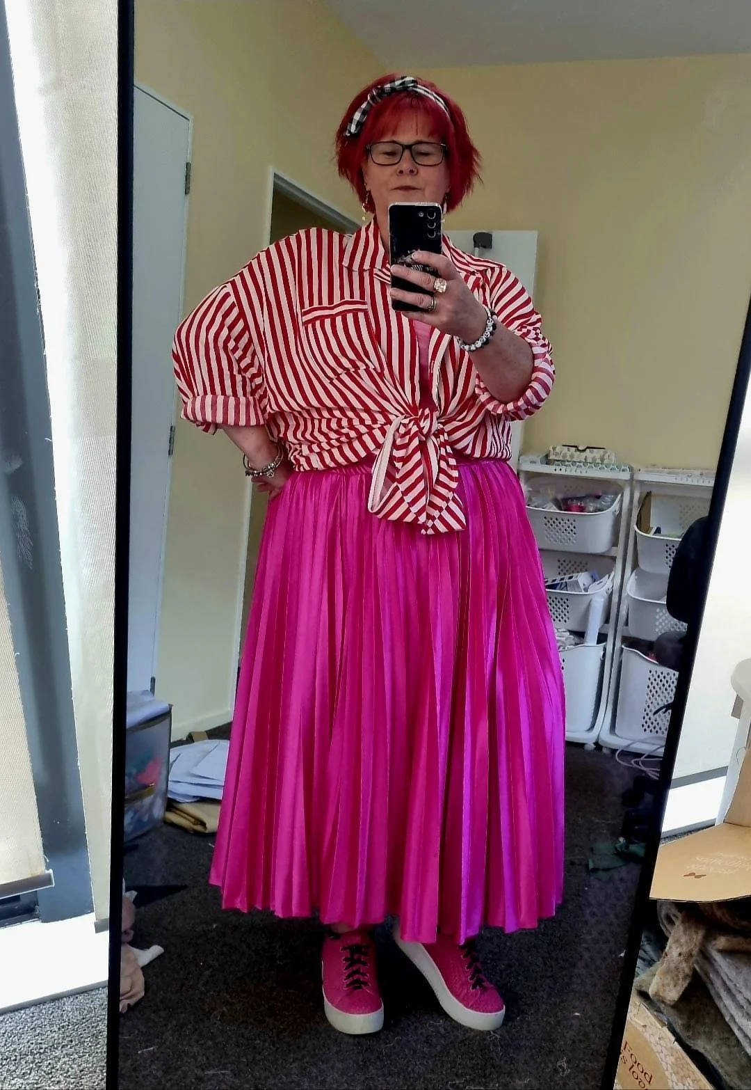A woman with short red hair, wearing glasses, a black and white headband, a red and white striped shirt tied at the waist, a long pink pleated skirt, and pink sneakers, taking a mirror selfie in a room with a yellow wall.