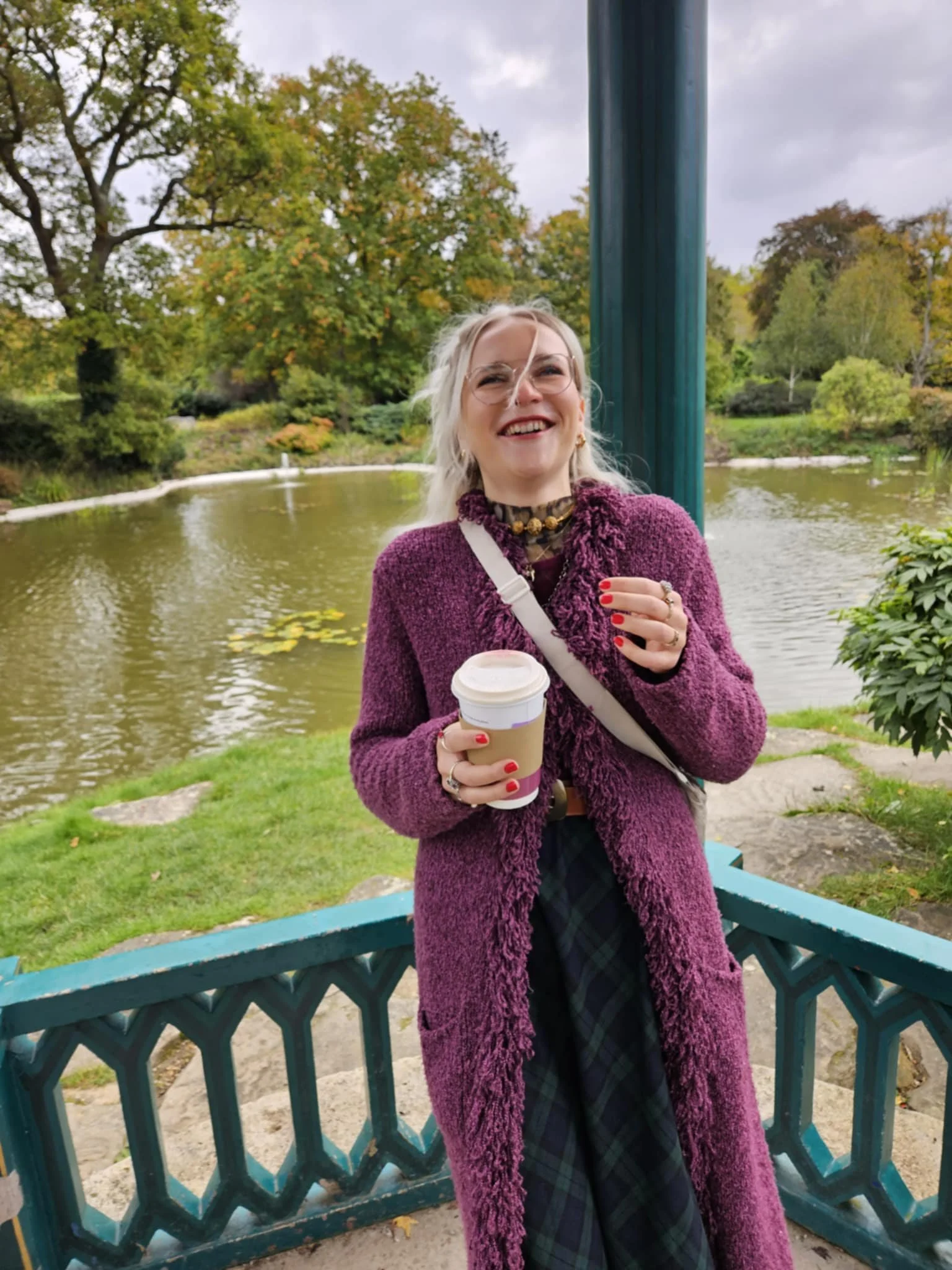 A woman with blonde hair and glasses smiling and holding a coffee cup, standing behind a teal park railing with trees and a pond in the background.