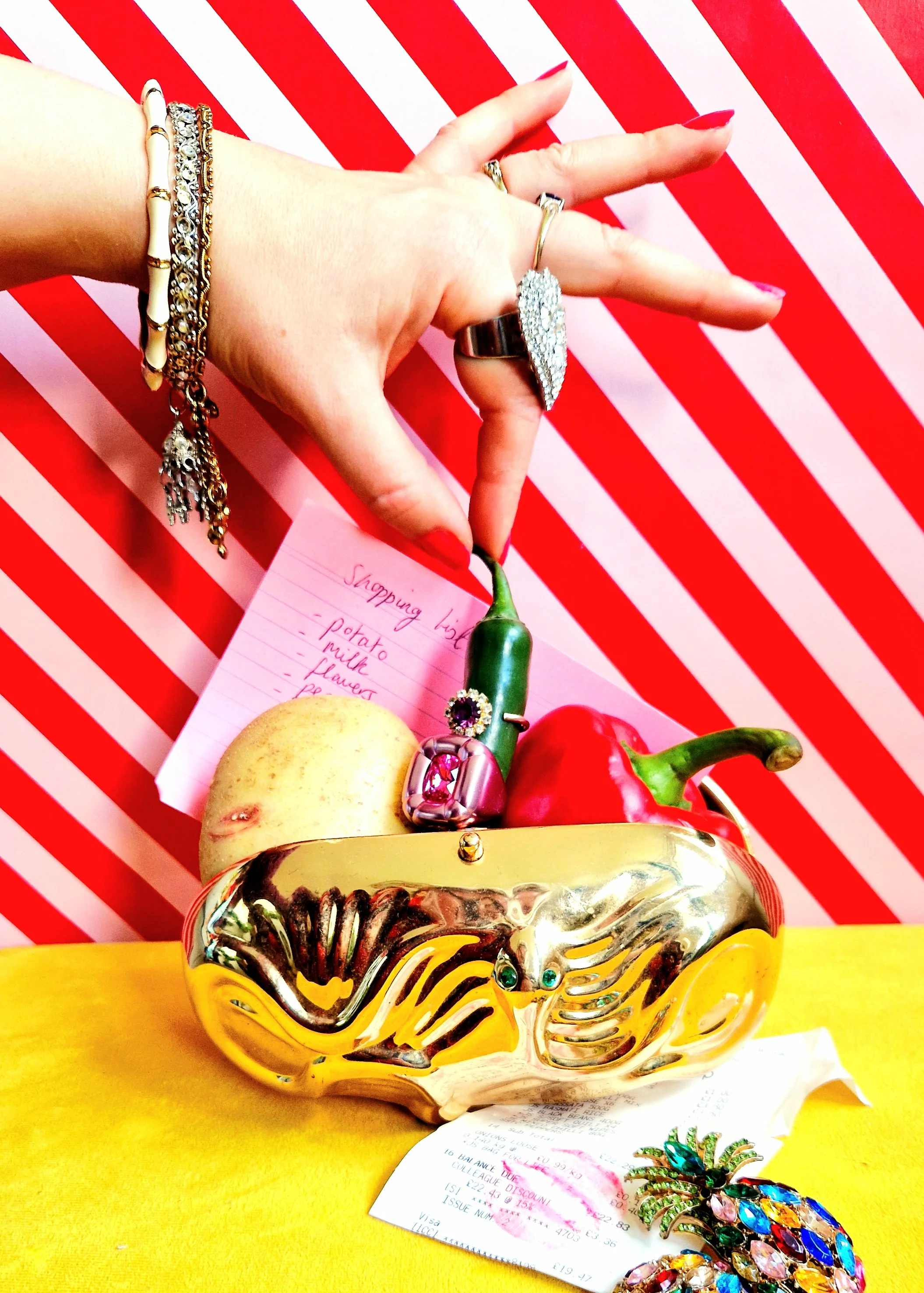 A hand with jewelry holding a green chili pepper over a decorative gold-colored bowl filled with fruit and jewelry, on a yellow surface with a red and white striped background.
